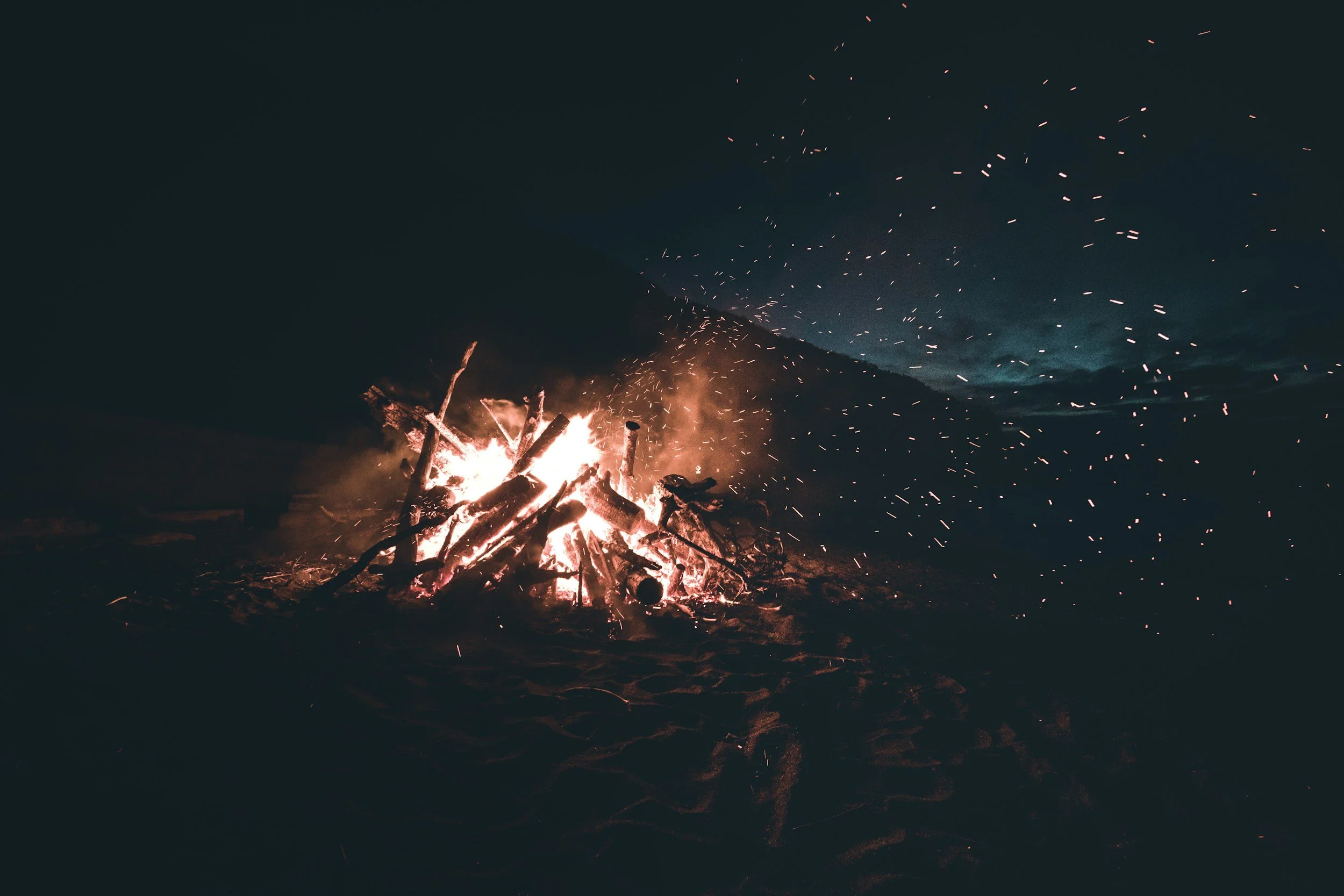 A campfire burning on the beach during nighttime with a dark sky and distant clouds.