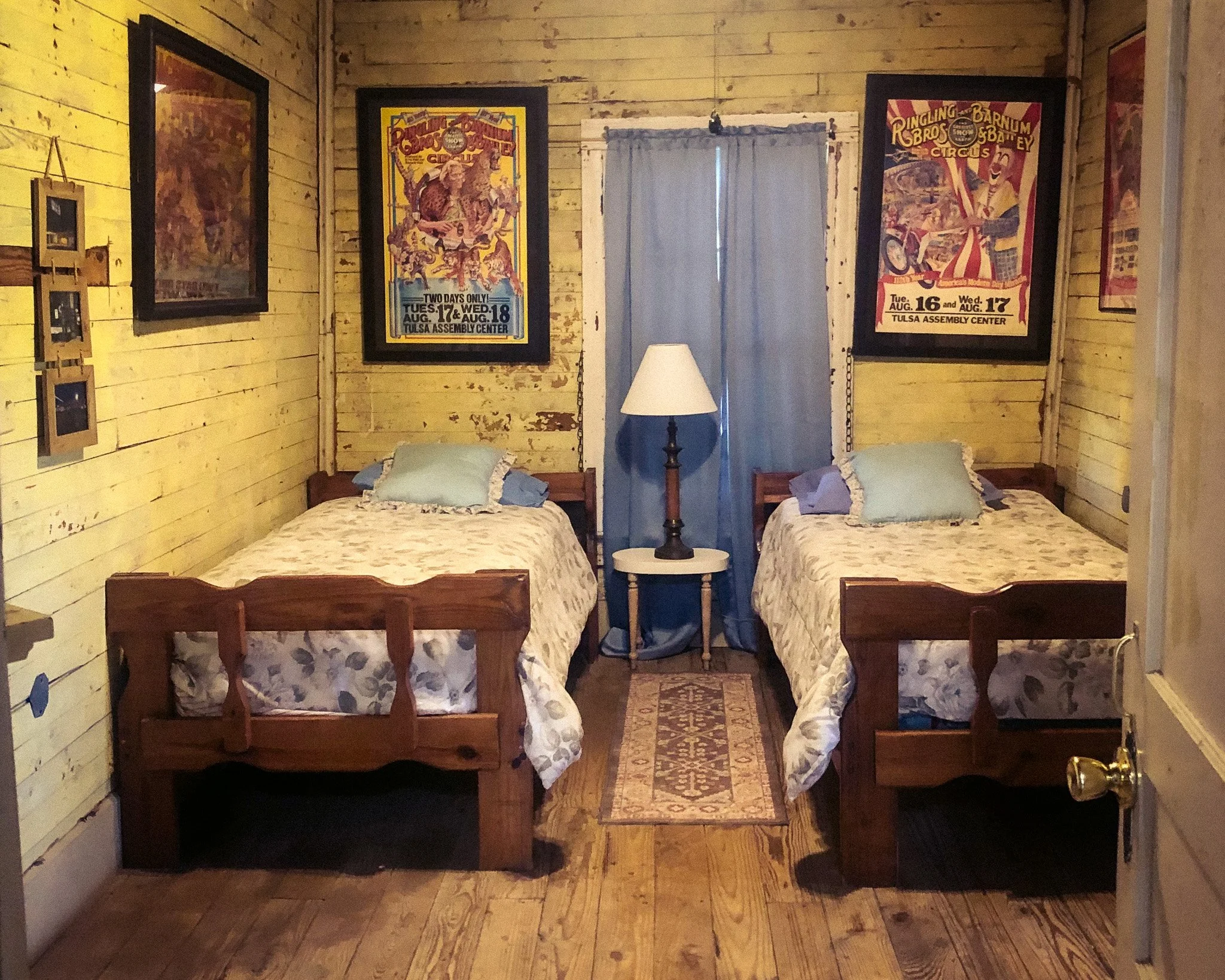 A rustic bedroom with two twin beds with white bedding and decorative pillows, a small circular braided rug on a wooden floor, a window with blue curtains, a ceiling fan, and a wooden wall with artwork.