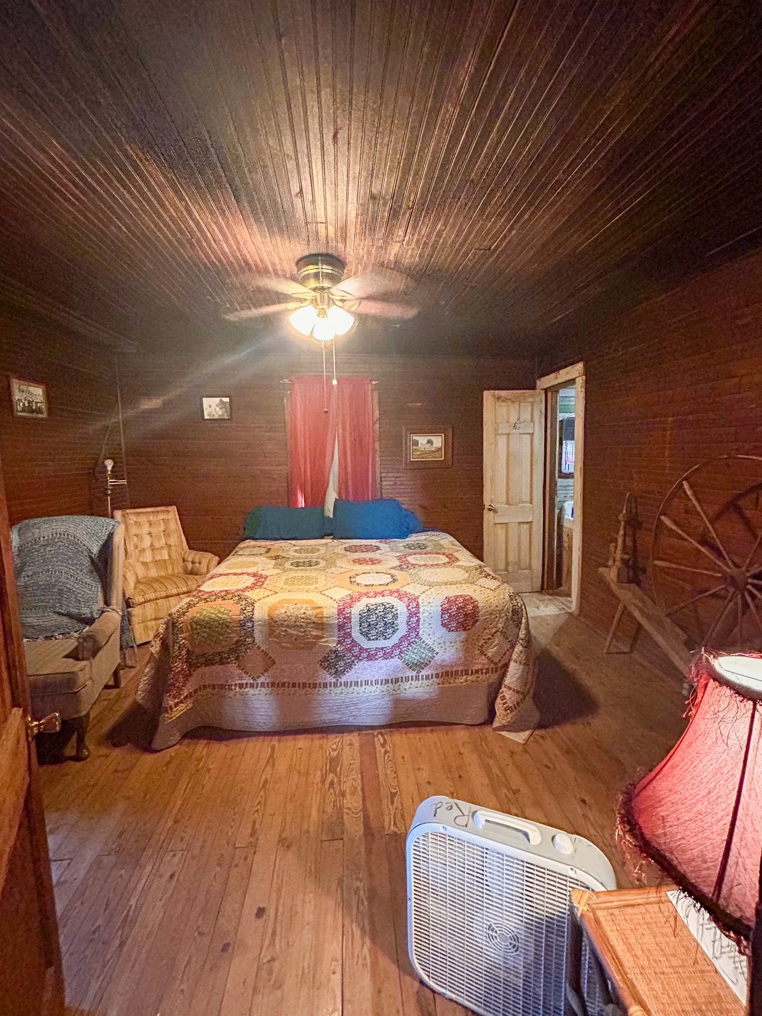 A cozy bedroom with wooden walls and ceiling, a bed with a colorful quilt, surrounded by chairs and a fan on the floor. There are framed pictures on the walls, a window with red curtains, and a partially open door.
