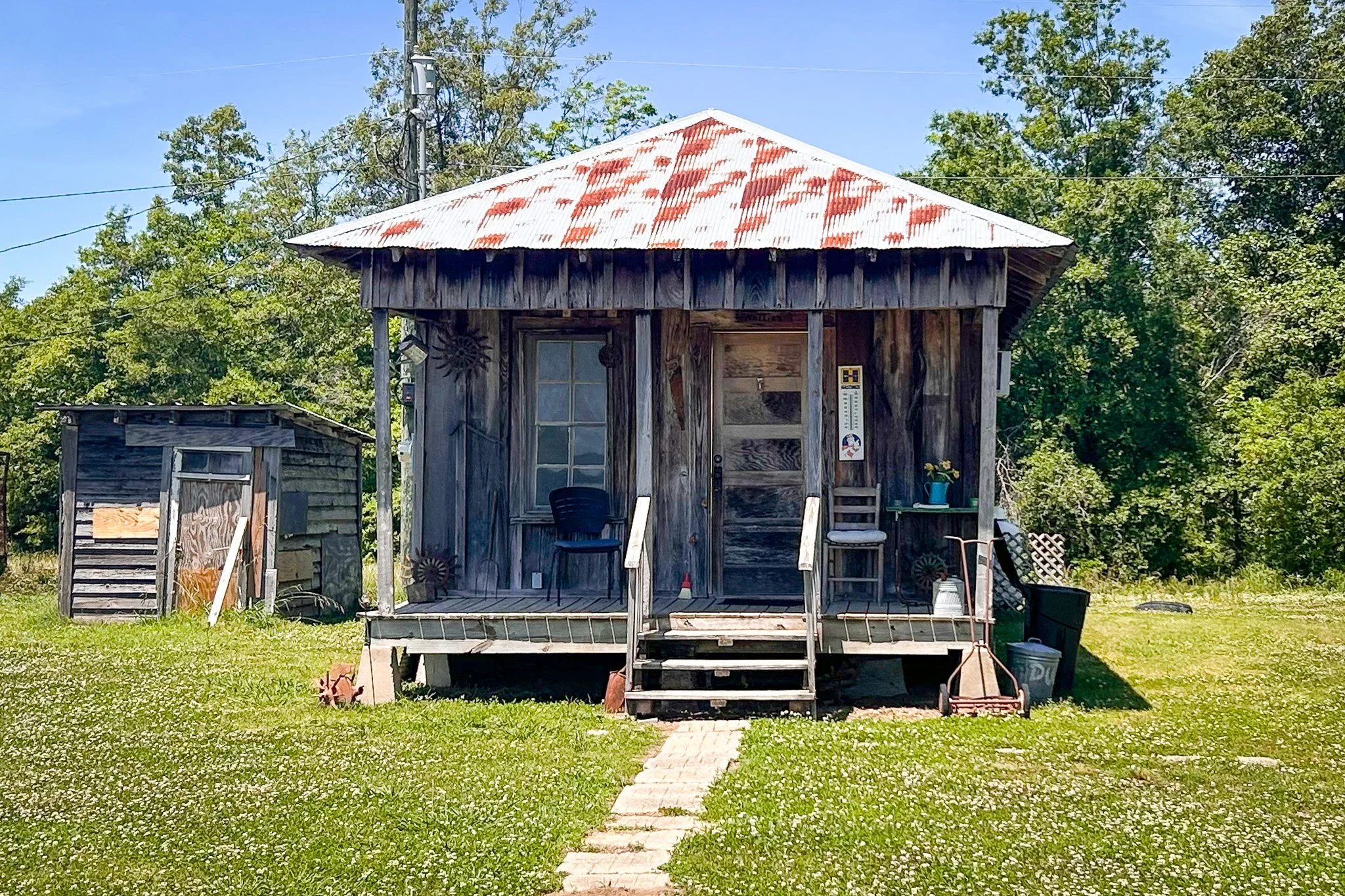 A small rustic wooden house with a corrugated metal roof, front porch, and steps leading up to it, surrounded by green grass and trees.
