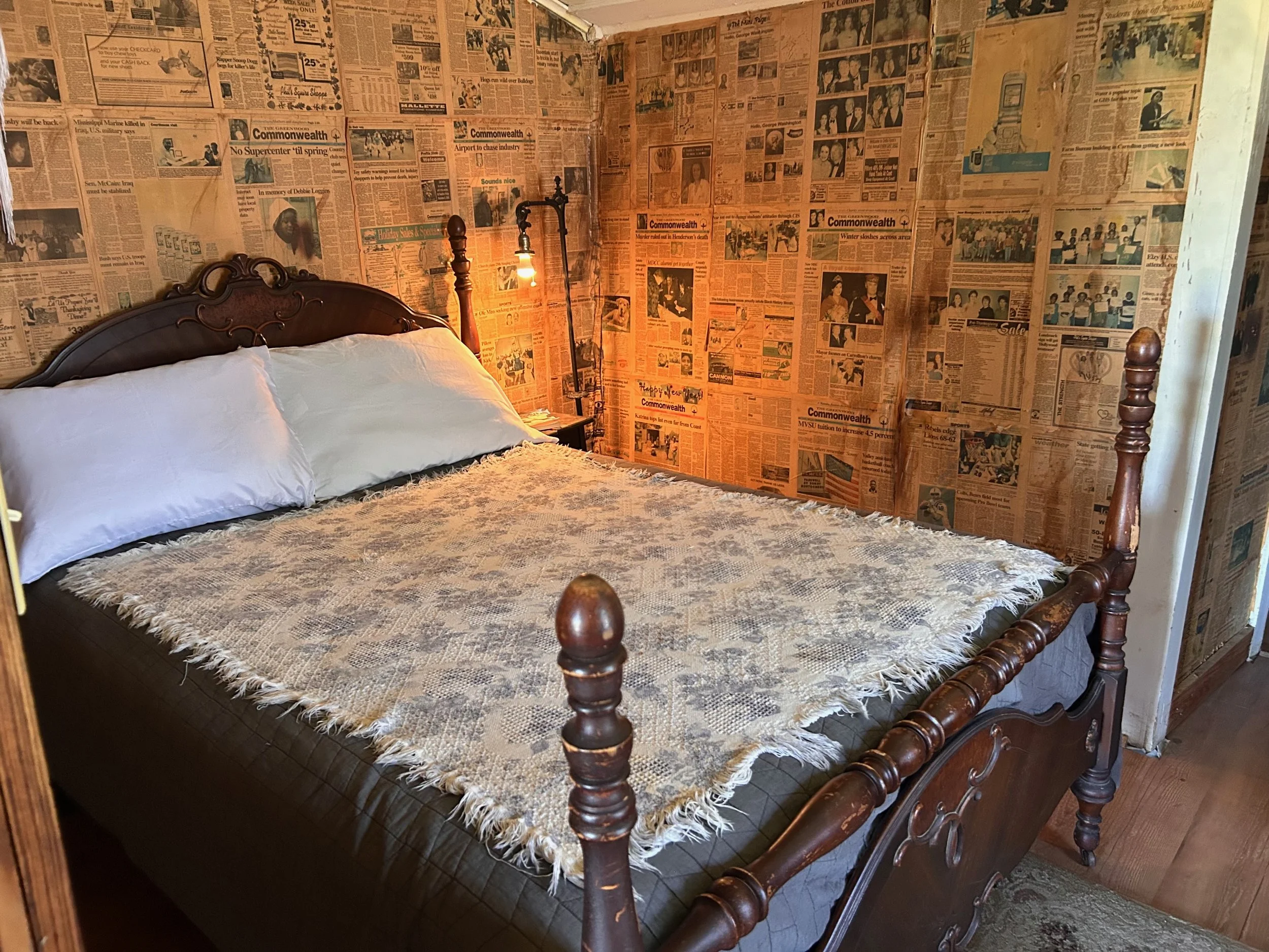 A bedroom with walls covered in old newspapers, a wooden bed frame, white pillows, a beige blanket, a small bedside table, and a vintage lamp with an exposed bulb.