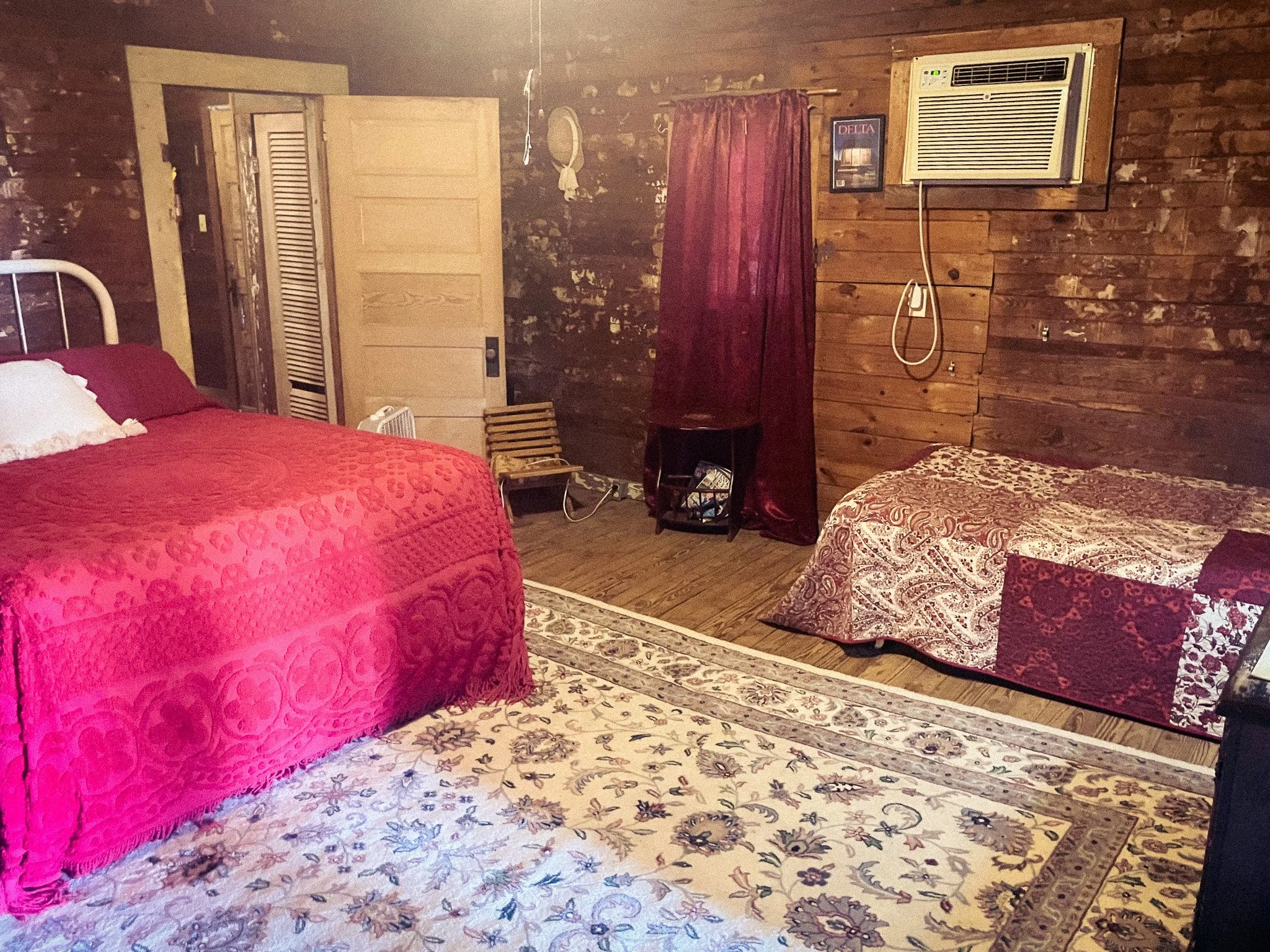 A bedroom with wooden walls and flooring, a bed with a red quilt, a paisley-patterned bedspread, a small chair, a vintage wooden chair, a window with red curtains, a wall-mounted air conditioning unit, framed artwork, and a patterned area rug.