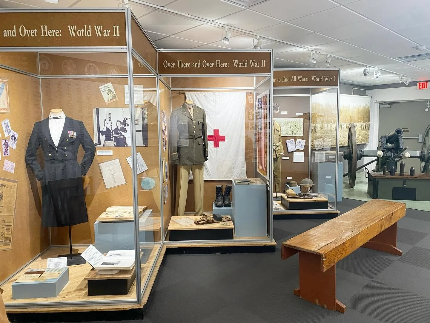 Museum exhibit displays World War II uniforms, photographs, and artifacts behind glass, with a large military machine in the background and a wooden bench in the foreground.