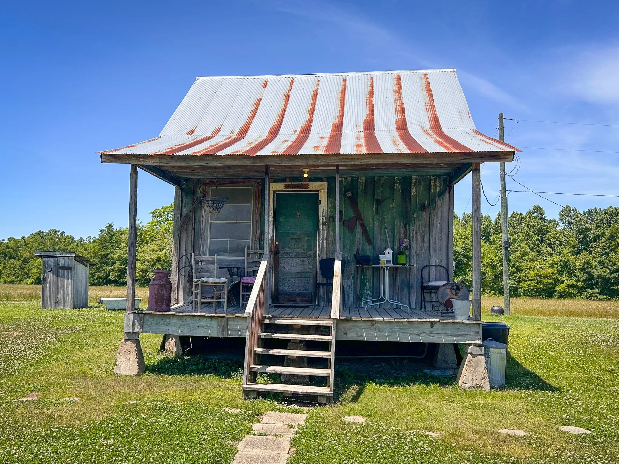 A small weathered wooden house with a rusted metal roof, a porch, and steps leading up to the front door, surrounded by green grass and trees under a blue sky.
