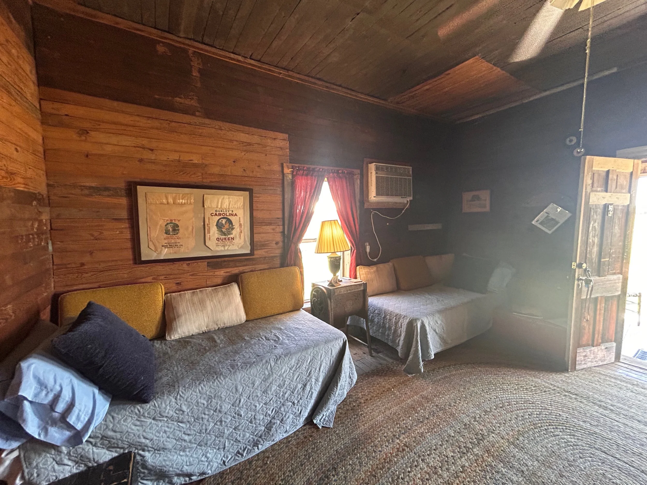 A cozy living room with wooden walls and ceiling, featuring a couch with pillows, a lamp, a framed poster on the wall, an air conditioning unit, and a window with red curtains, illuminated by natural light.