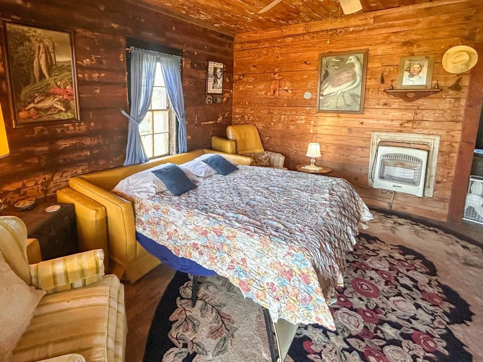 A rustic bedroom with wooden paneled walls and ceiling, featuring a large bed with a floral quilt, a yellow upholstered headboard, and matching armchair. Decor includes framed artwork of birds and a portrait, table lamps, a ceiling fan, an elephant-shaped heater, and a window with blue curtains.