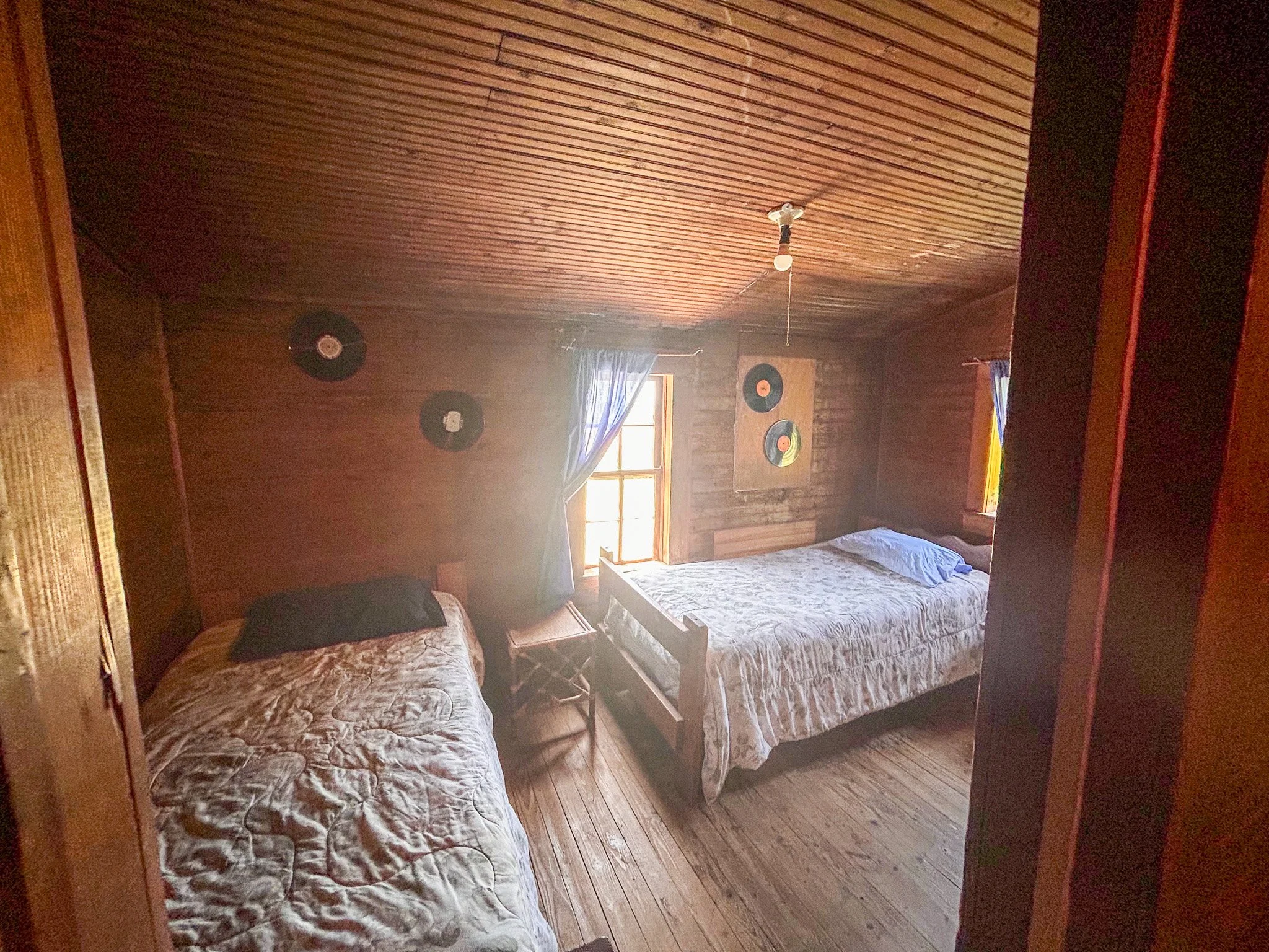 A cozy bedroom with wooden walls and ceiling, two beds with patterned bedspreads, a window with curtains, and vintage vinyl records on the wall.