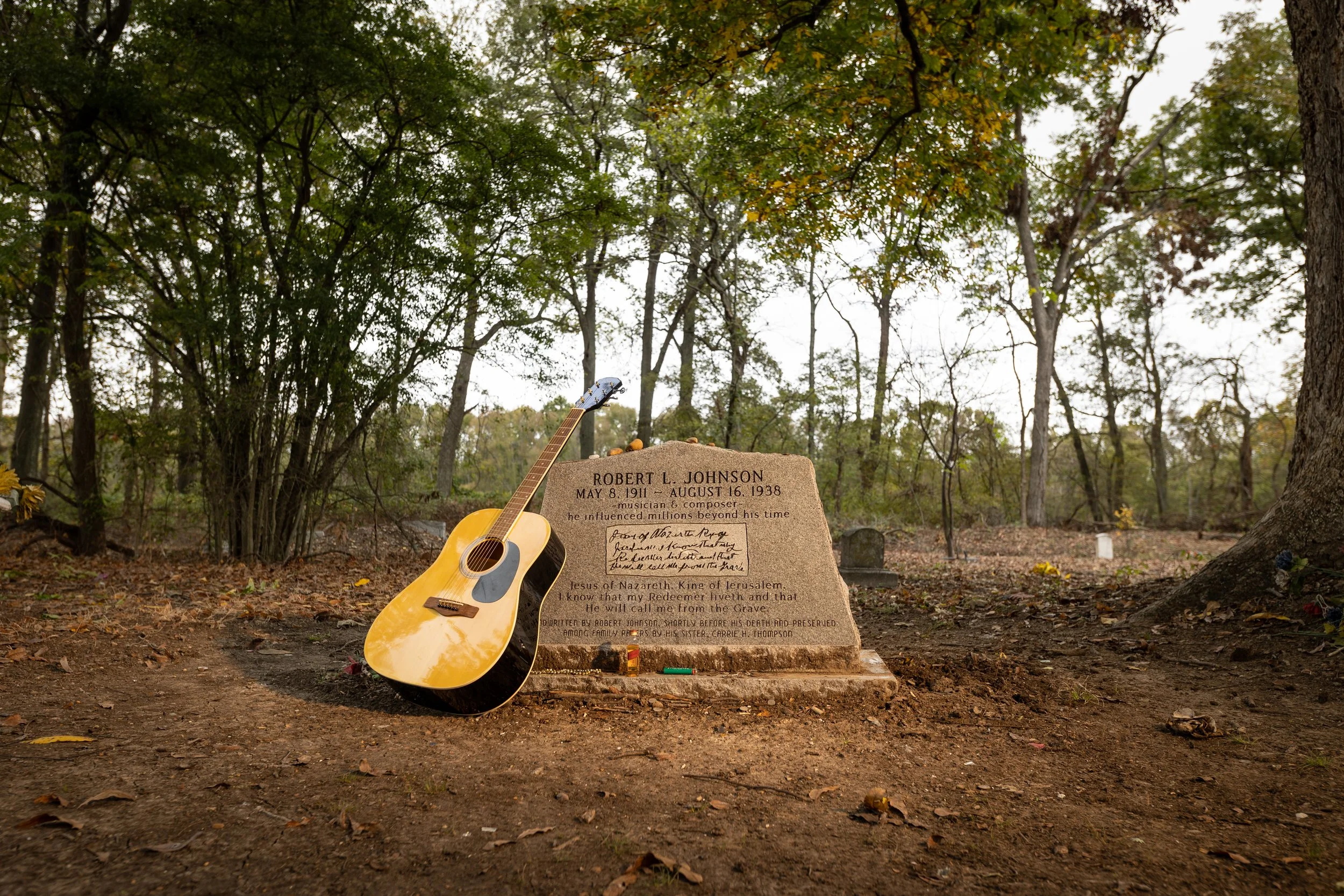 Gravestone with a guitar leaning against it in a cemetery surrounded by trees.