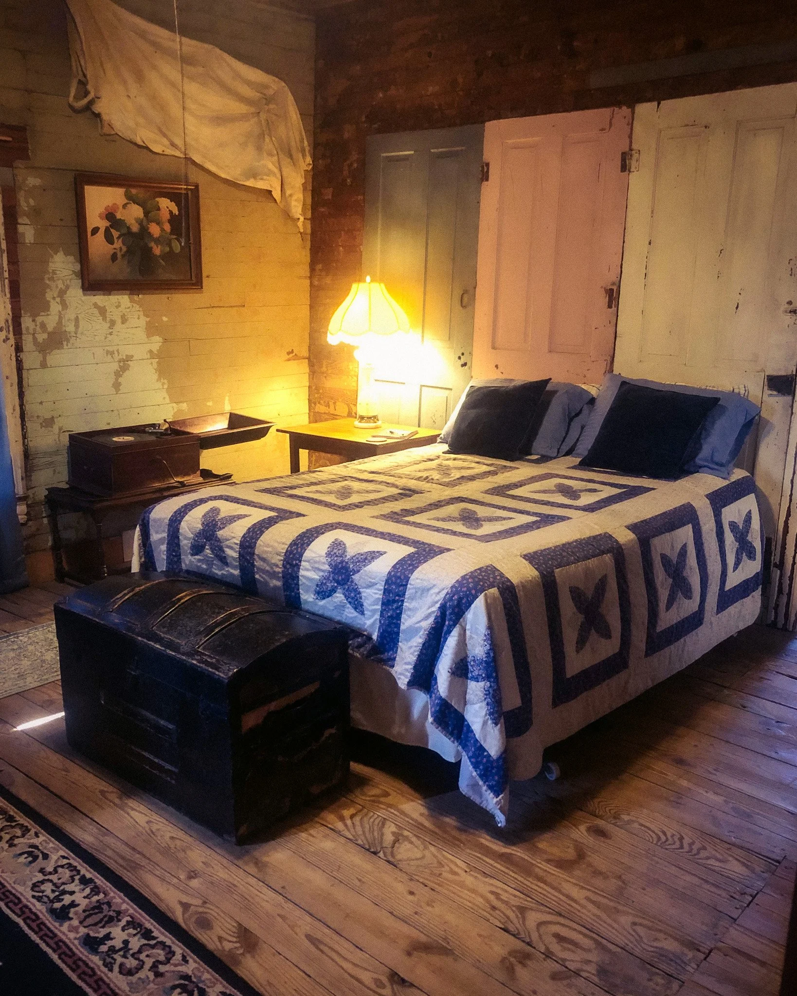 A rustic bedroom with wooden walls and ceiling, featuring a bed with a blue and white patterned quilt, a wooden nightstand with a lamp, a window with blue curtains, and a ceiling fan.