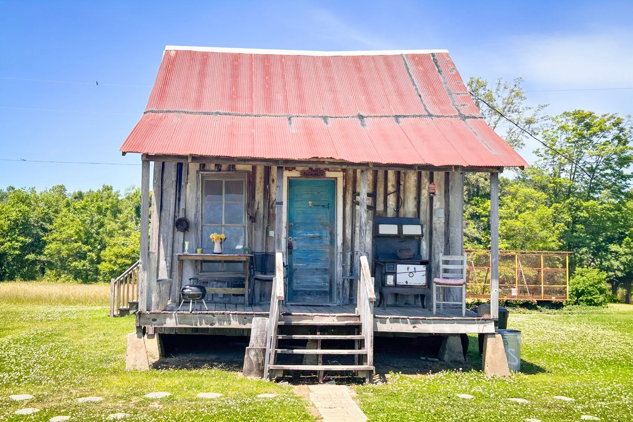 An old wooden house with a red rusted metal roof, weathered blue front door, and porch with stairs, surrounded by green trees and open field under a clear blue sky.