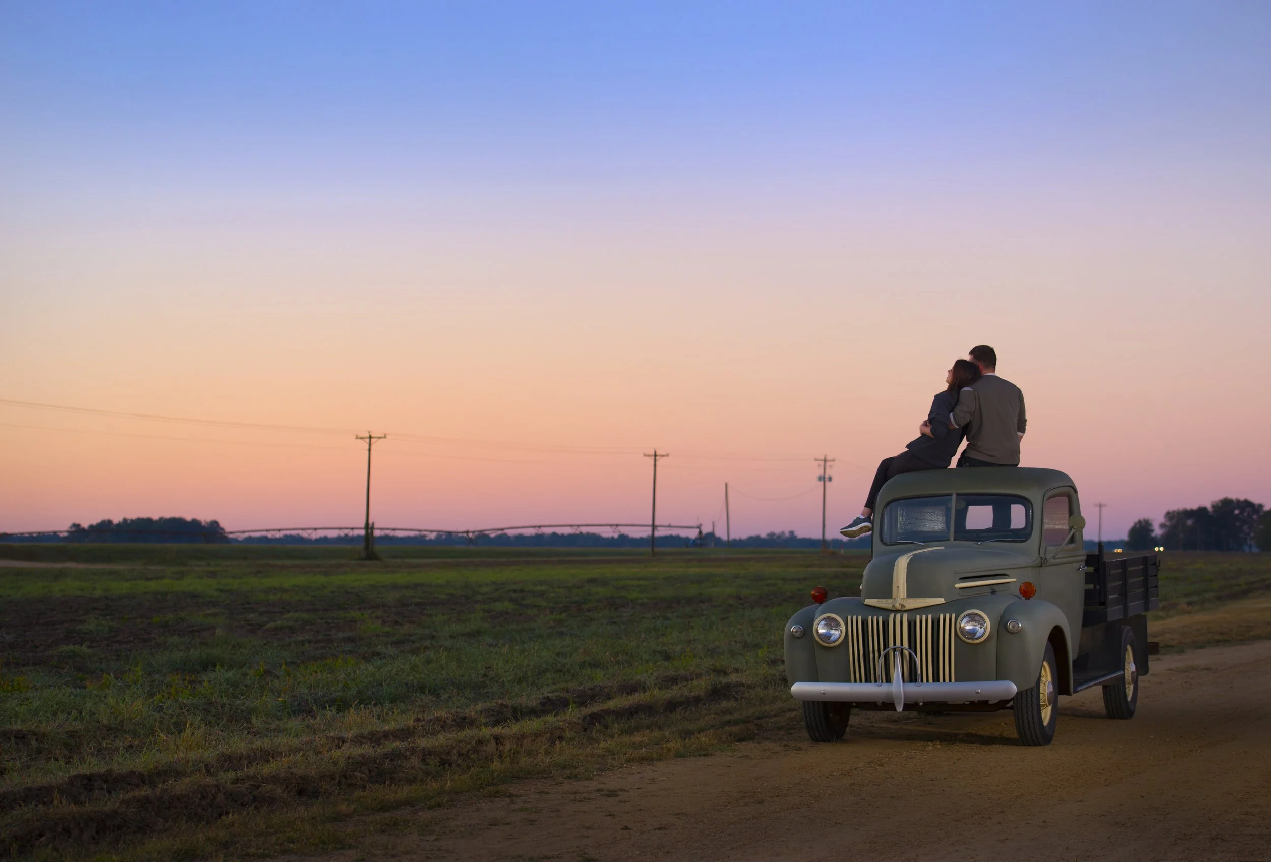 A couple sitting together on top of an old truck in a rural field during sunset, with power lines and trees in the background, and a colorful sky.