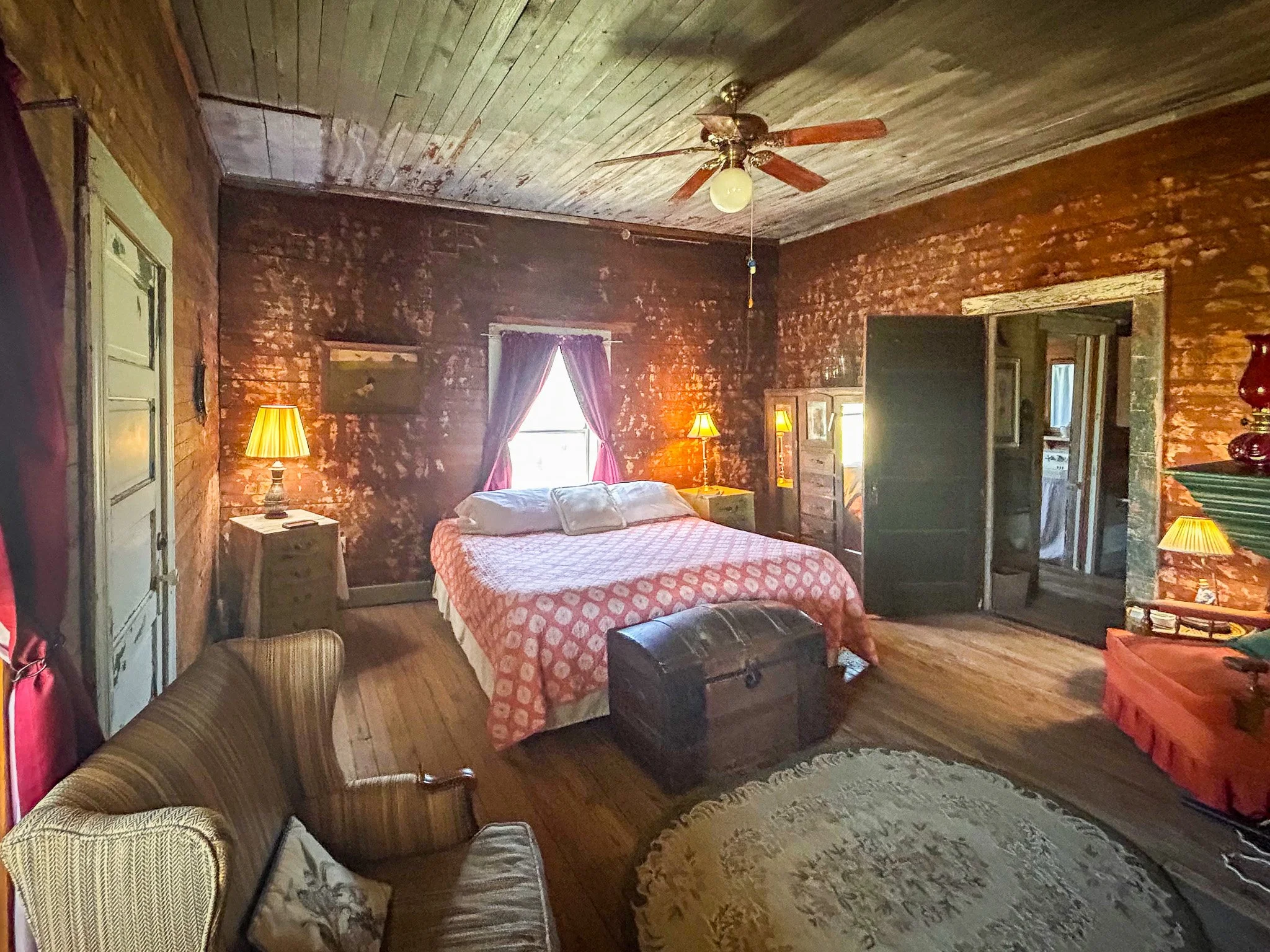 A rustic bedroom with wooden walls and ceiling, featuring a bed with a polka dot blanket, illuminated by two lamps on nightstands, and decorated with vintage furniture and framed artwork. There is a window with curtains, and a ceiling fan with a light.