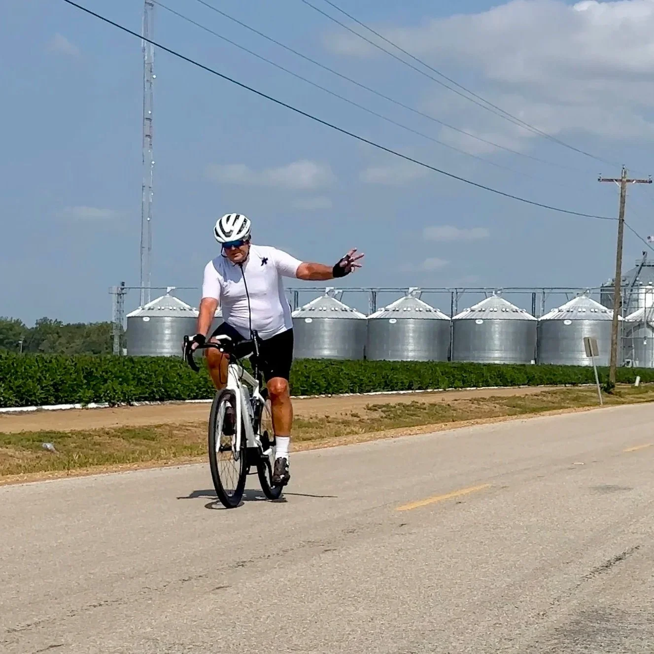 Man in cycling gear riding a bicycle on rural road, farm silos and utility poles in background.