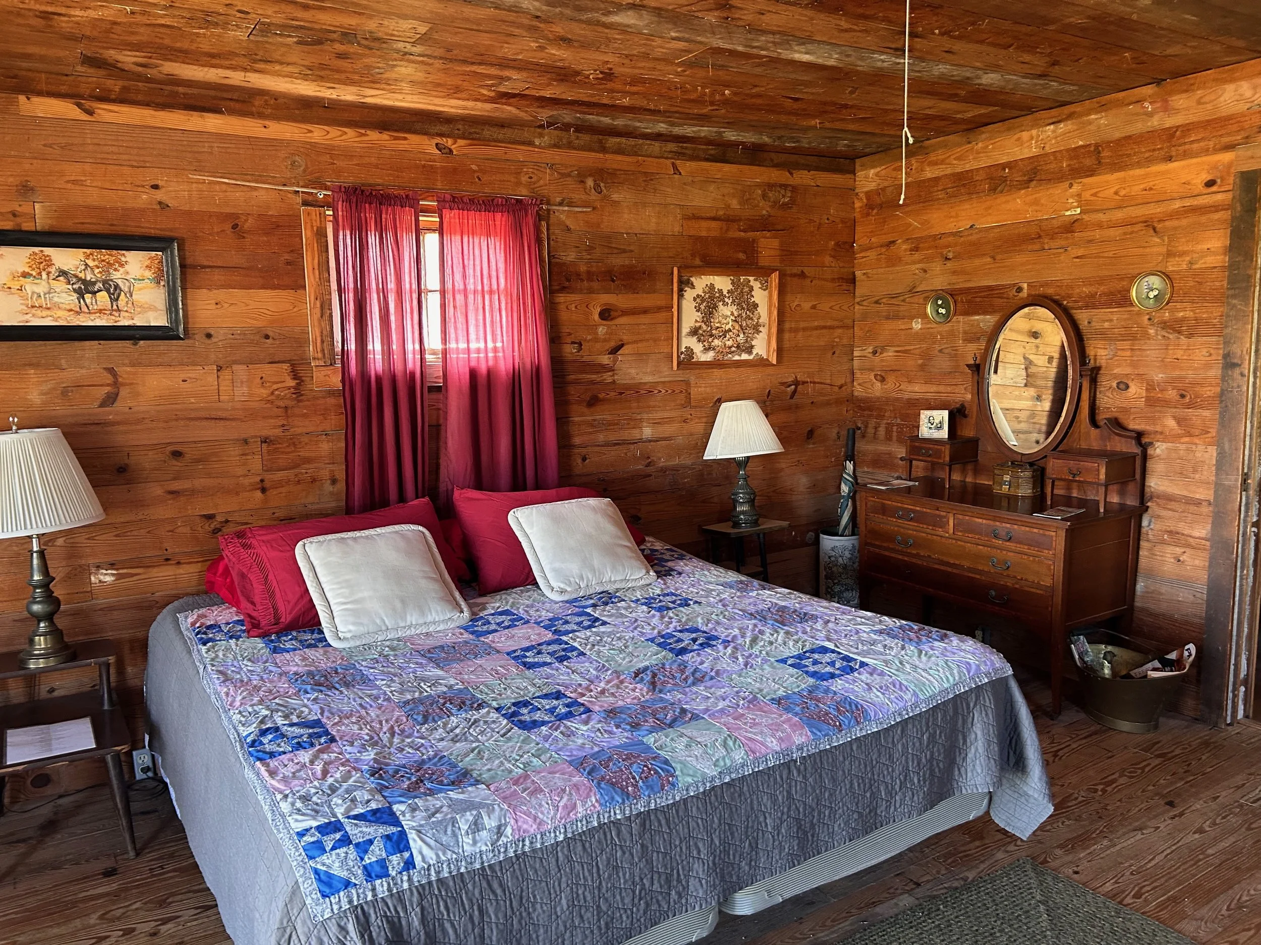 A rustic bedroom with wooden walls and ceiling, featuring a bed with a colorful patchwork quilt and white and red pillows, two bedside lamps, a mirror on a dresser, and framed artwork of animals and a tree on the walls. A window with red curtains is behind the bed.