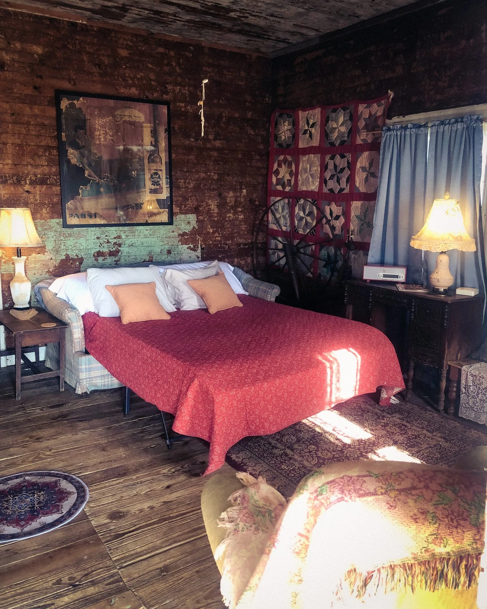 A cozy living room with wood-paneled walls and a wooden ceiling. There is a blue sofa with two pillows, a small wooden coffee table, and a patterned area rug. Decor items include a table lamp, a framed mirror or picture on the wall, and a red quilt or blanket hanging on a wagon wheel. A window with light blue curtains overlooks the room, and part of the kitchen with a microwave and counter is visible through an open doorway.