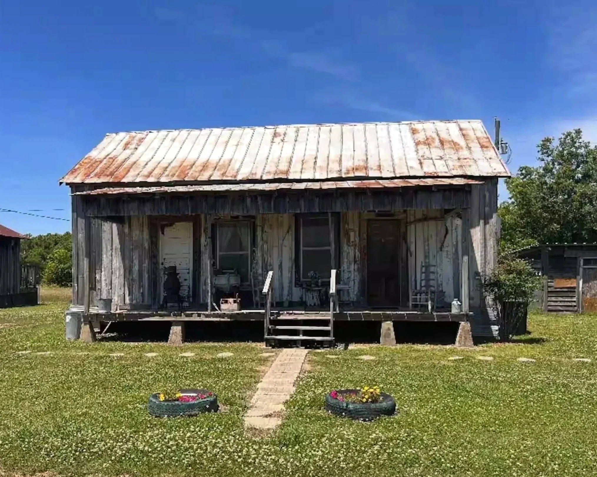 A weathered wooden house with a rusted metal roof, front steps, two flower pots on the grass, a grassy yard, and trees in the background under a clear blue sky.