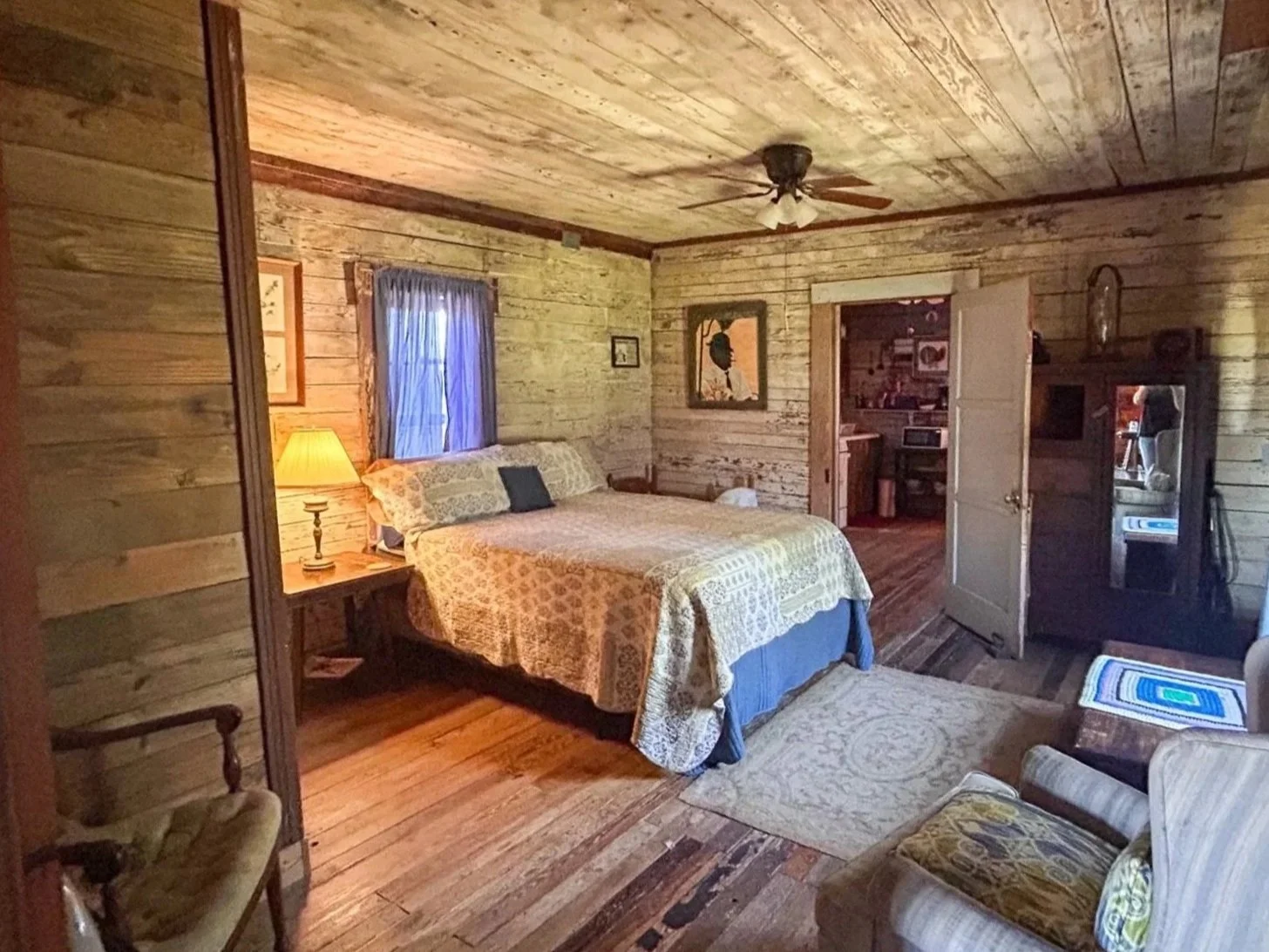 A cozy bedroom with wood-paneled walls and ceiling, a bed with patterned bedding, a lamp on a bedside table, a small window with blue curtains, a ceiling fan, a sitting area with a striped armchair and a floral armchair, a black cabinet, and an open door leading into another room.