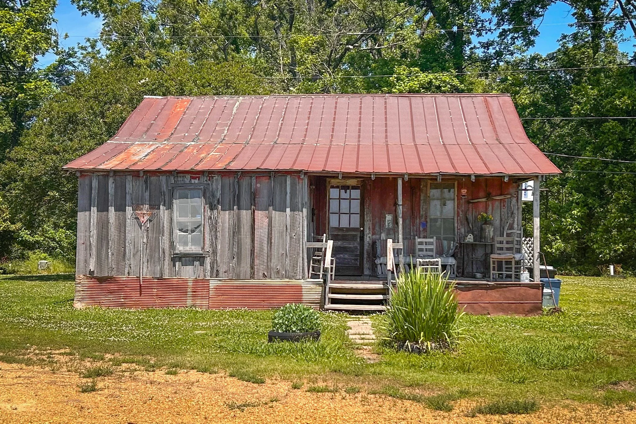 Old weathered house with a rusted metal roof, surrounded by greenery and trees, with a small porch and steps at the entrance.