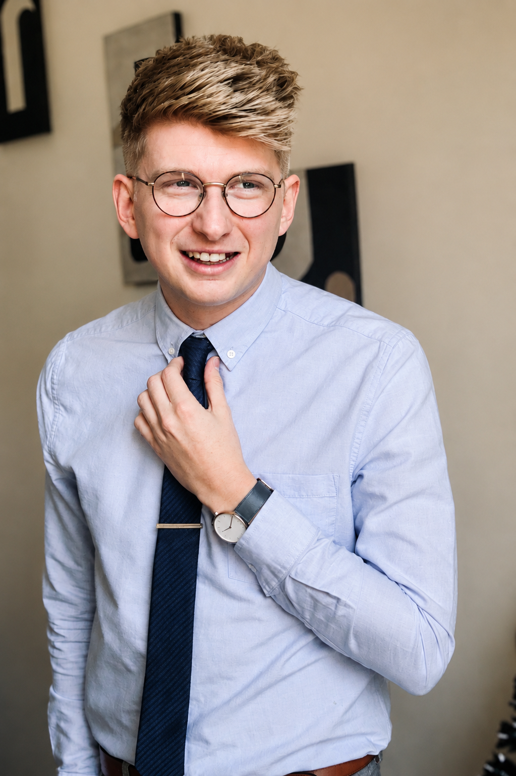 A young man with light brown hair and glasses, wearing a light blue shirt and dark blue tie, is adjusting his tie with his left hand and smiling at the camera.