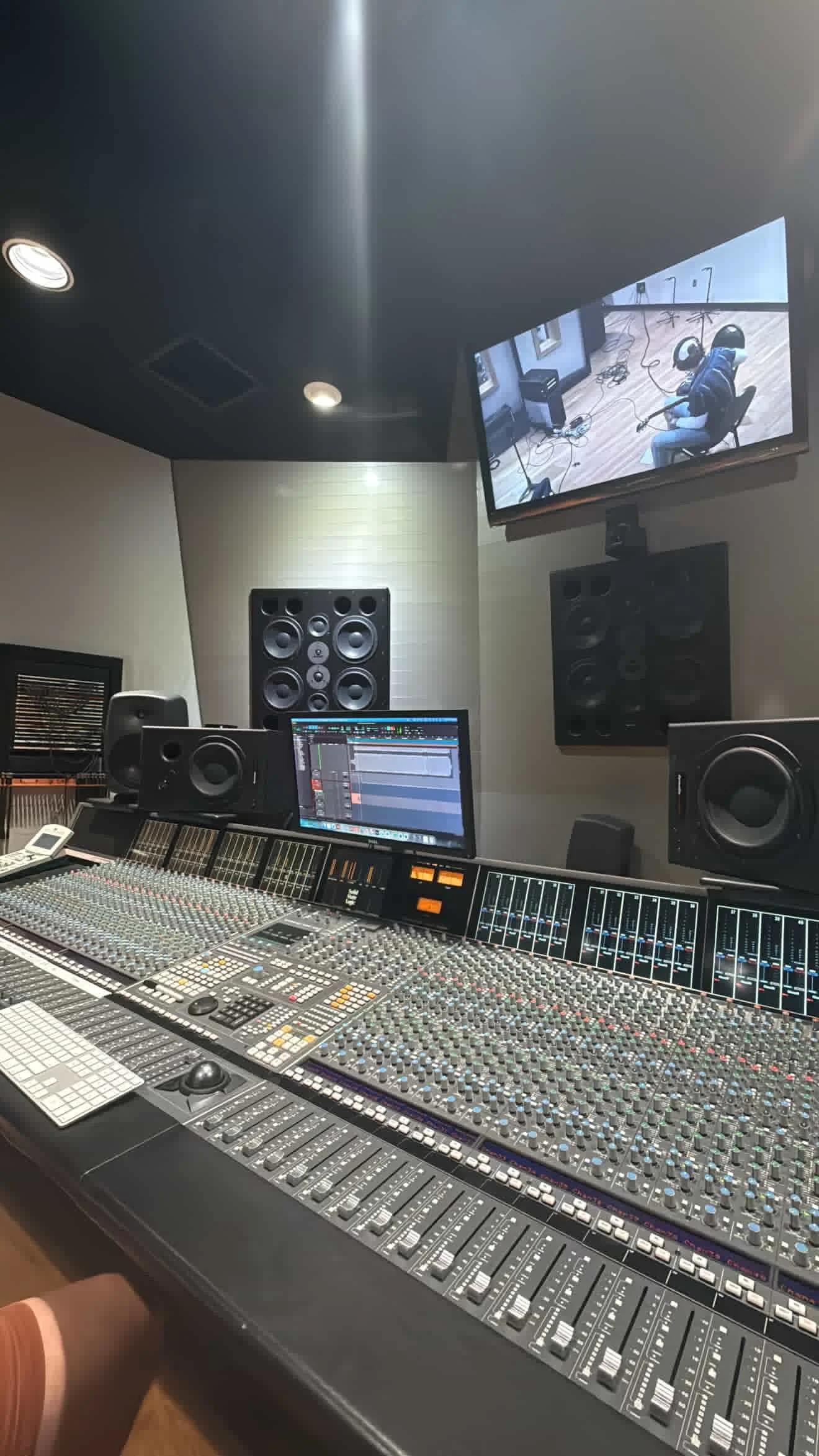 A professional recording studio with a large mixing console, studio monitors, and a computer screen, along with a wall-mounted TV displaying a person working in the studio.