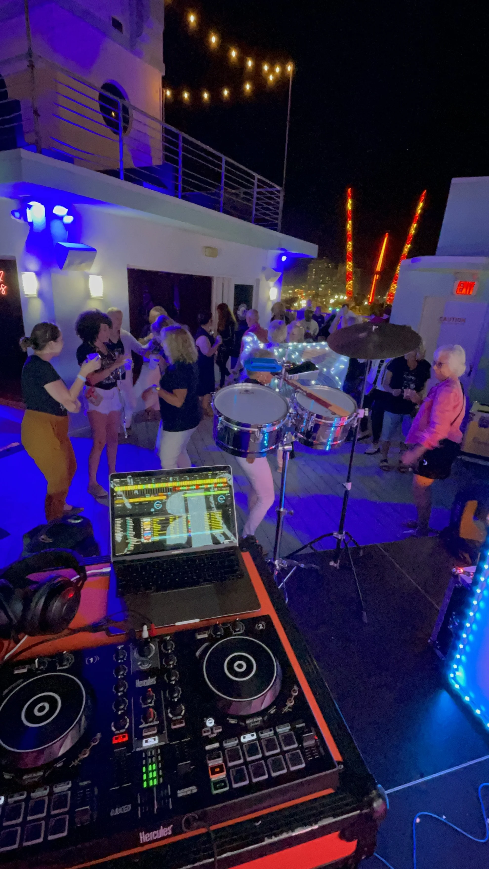 Nighttime rooftop party with people dancing and socializing, DJ equipment in foreground, colorful string lights, city lights in distance.