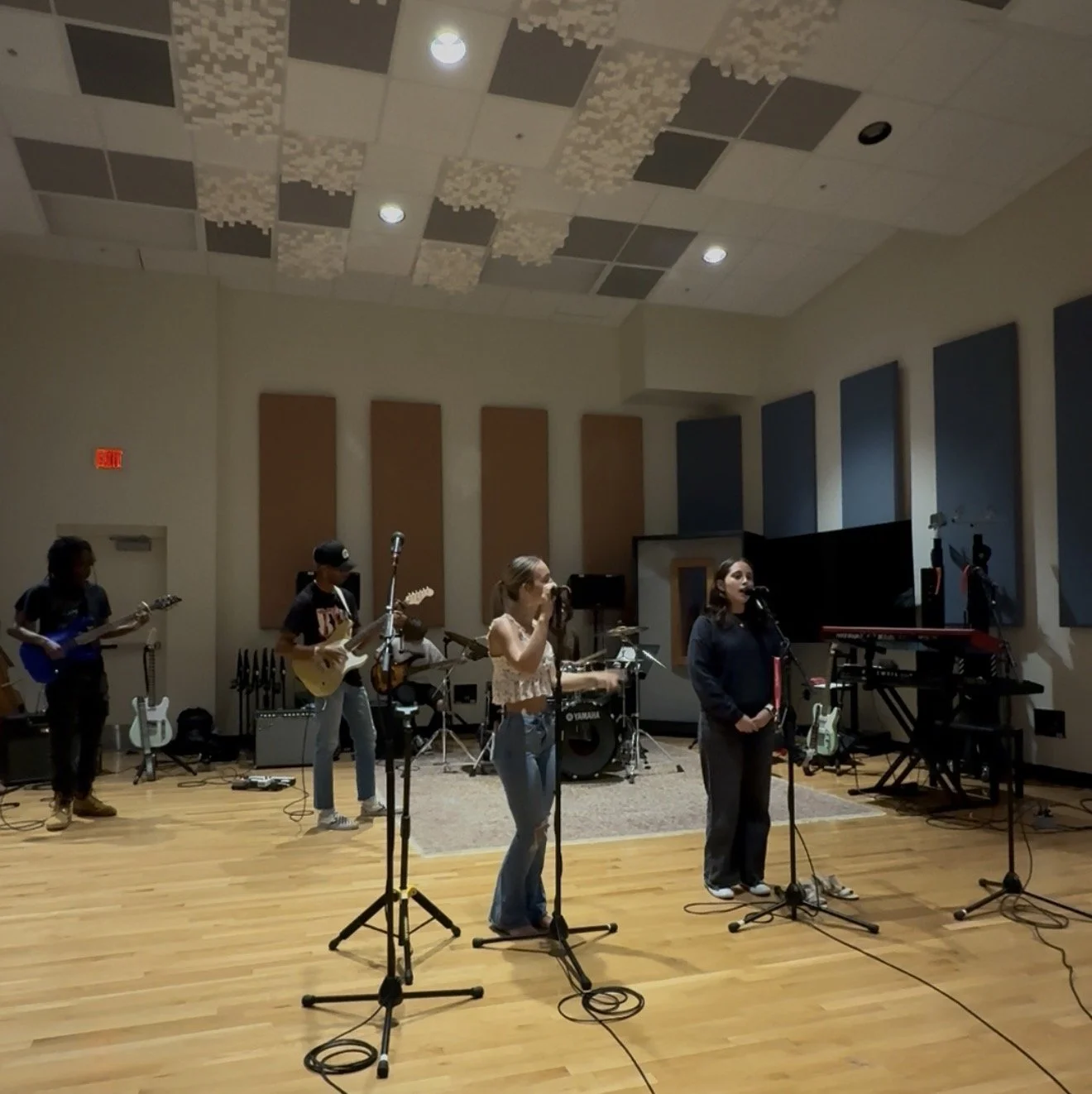 Group of five musicians performing in a recording studio, with guitars, drum set, keyboards, microphones, and soundproofing on the walls.