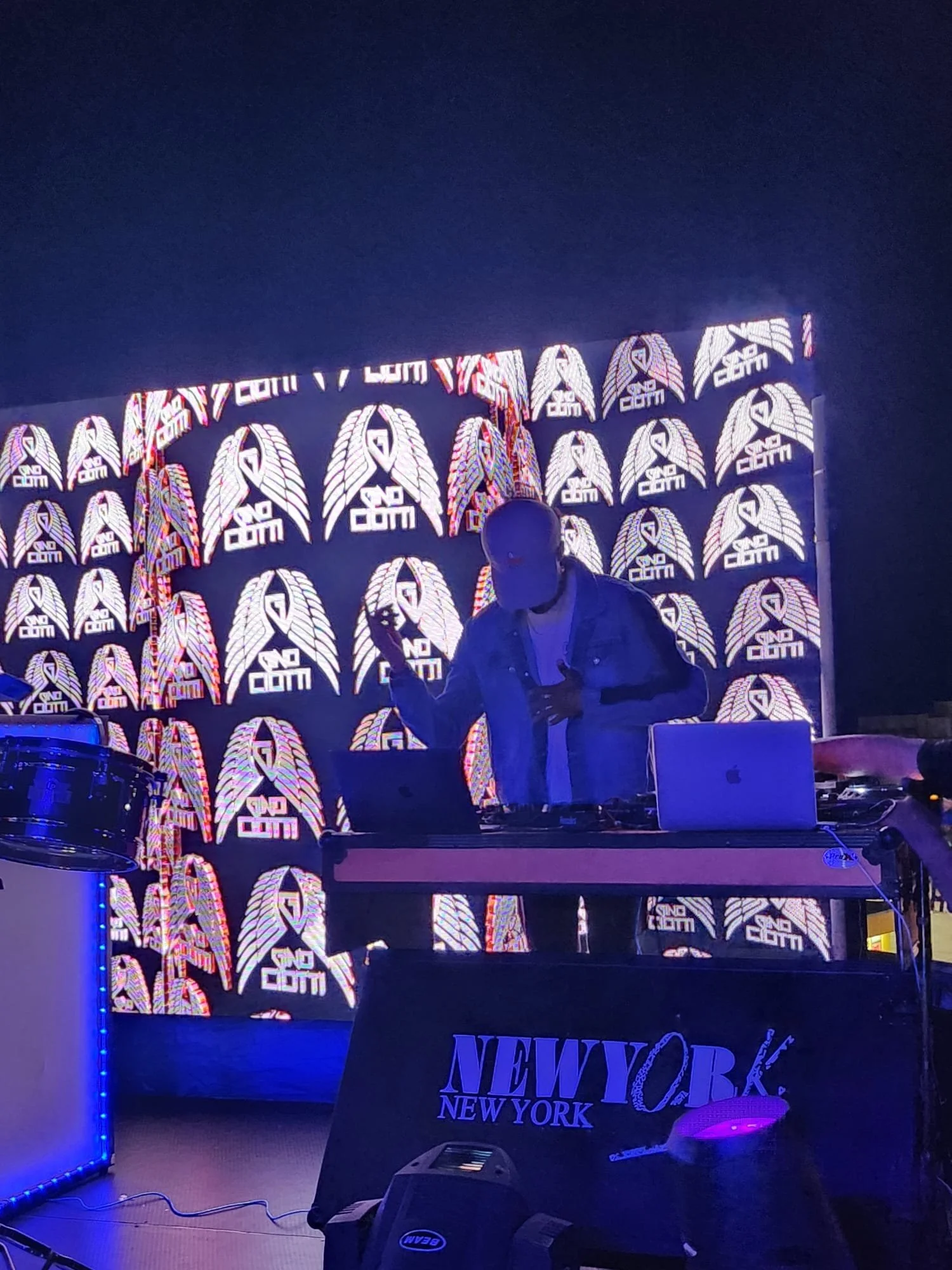 A person performing as a DJ at a nightclub or music event, standing behind DJ equipment with a laptop. The background has a wall decorated with multiple illuminated logos and designs, including the words 'sound.com' and a winged emblem, set against a dark blue atmosphere with blue lighting accents.