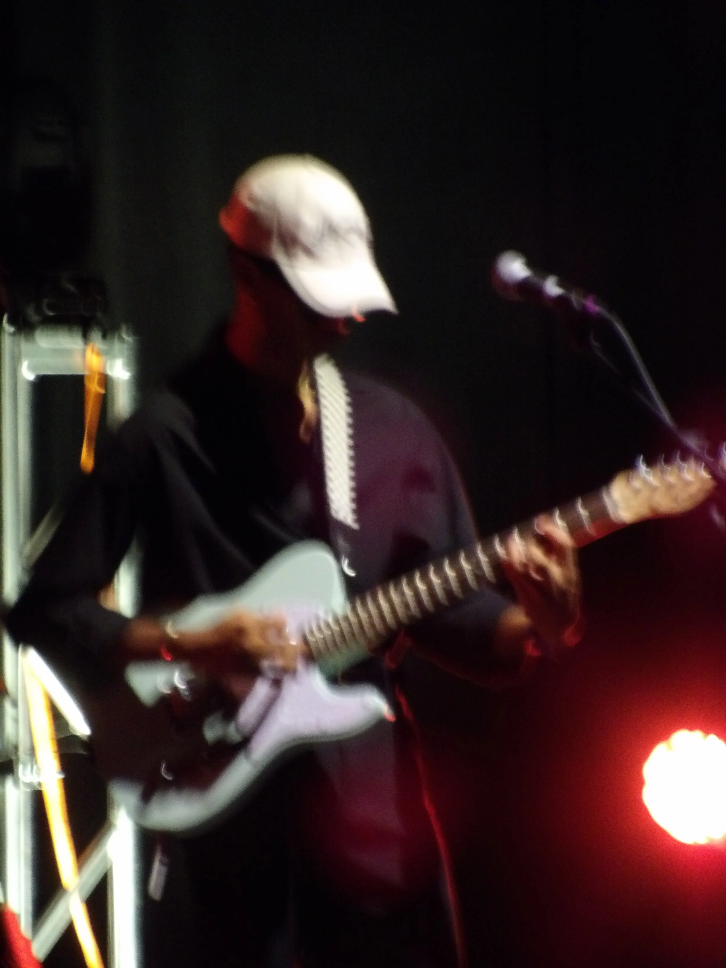 A person wearing a white baseball cap playing an electric guitar on stage with a microphone nearby, against a dark background with some stage lights.