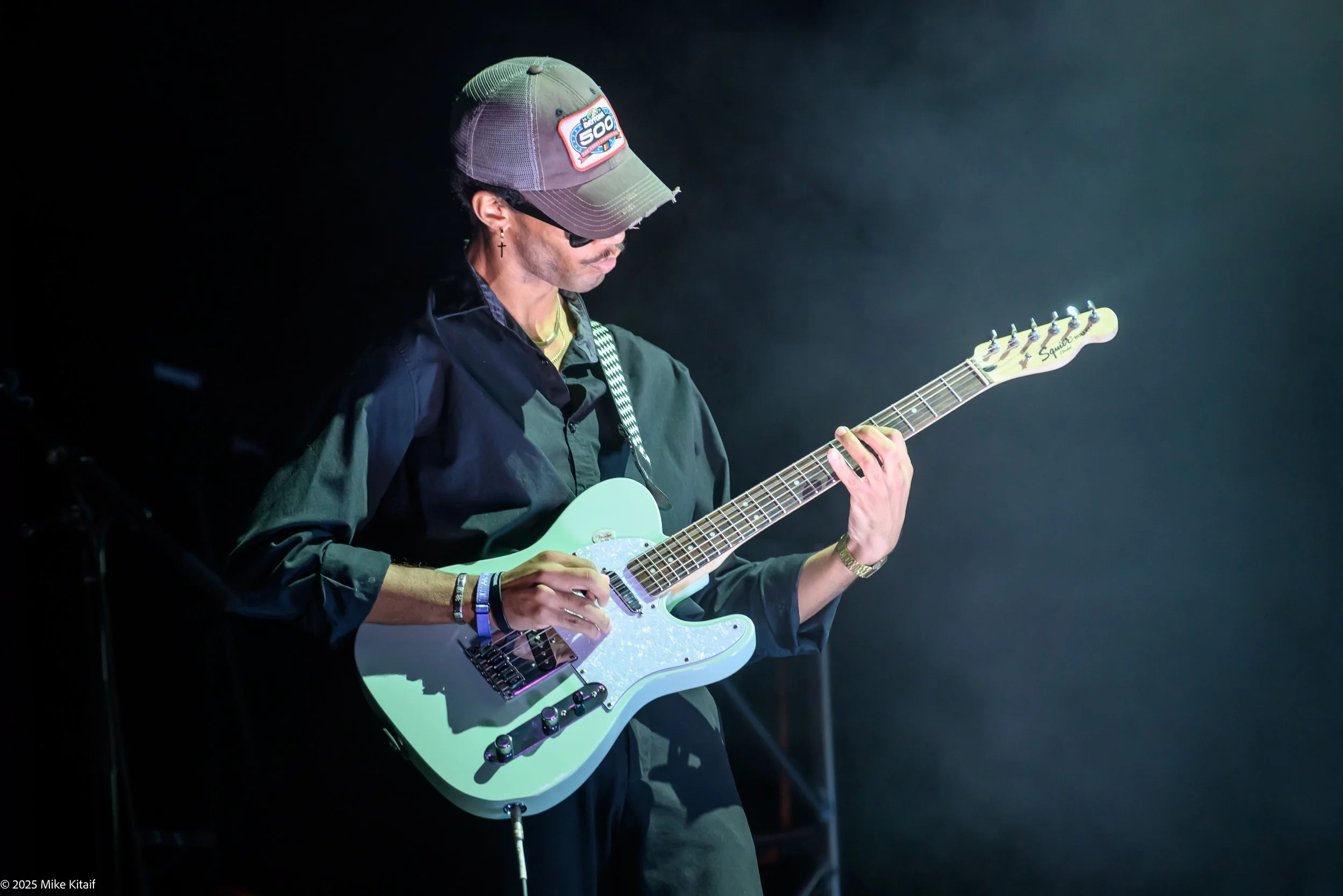A man wearing a cap with a patch reading 'NASCAR 500' on it, sunglasses, a cross earring, and several bracelets, playing a light green electric guitar on stage with a dark background.