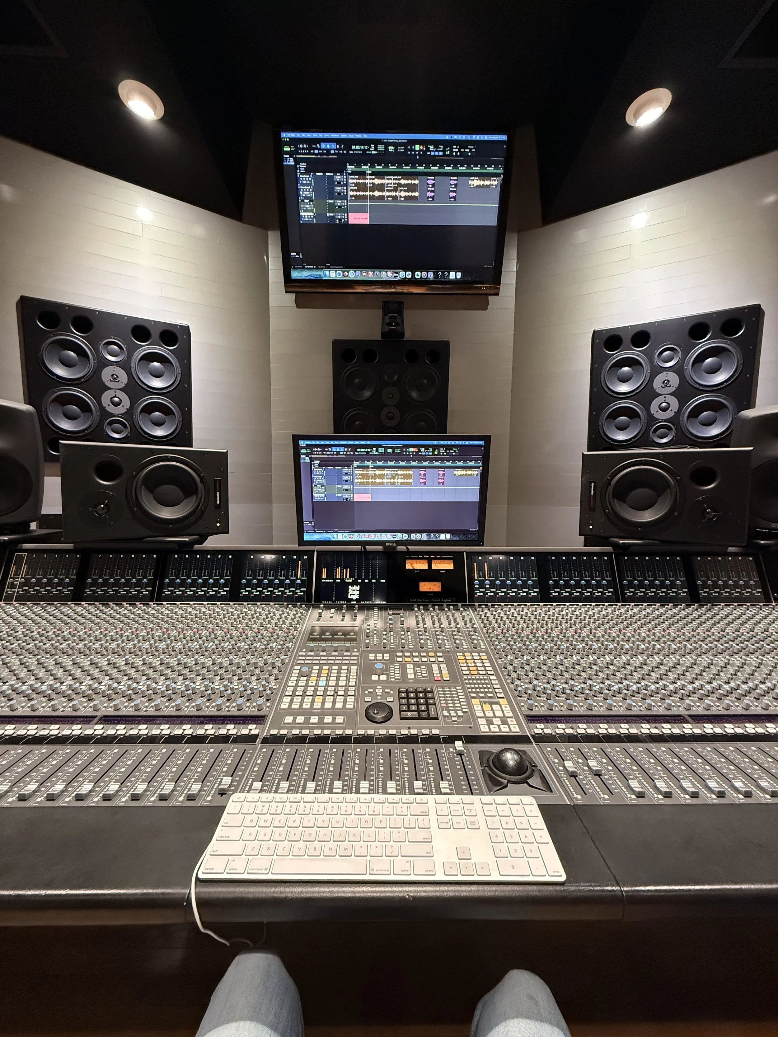 Recording studio mixing console and computer screens with audio editing software, speakers, and sound equipment.