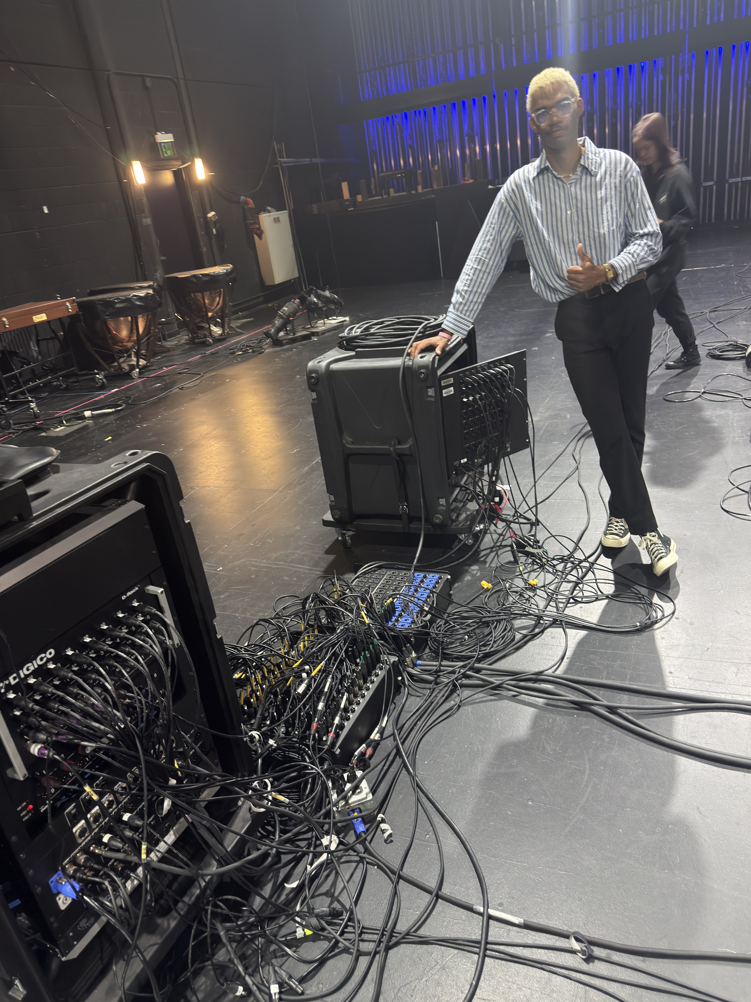 A man in a striped shirt and glasses standing on a stage with sound equipment, cables, and a dark backdrop, giving a thumbs-up.