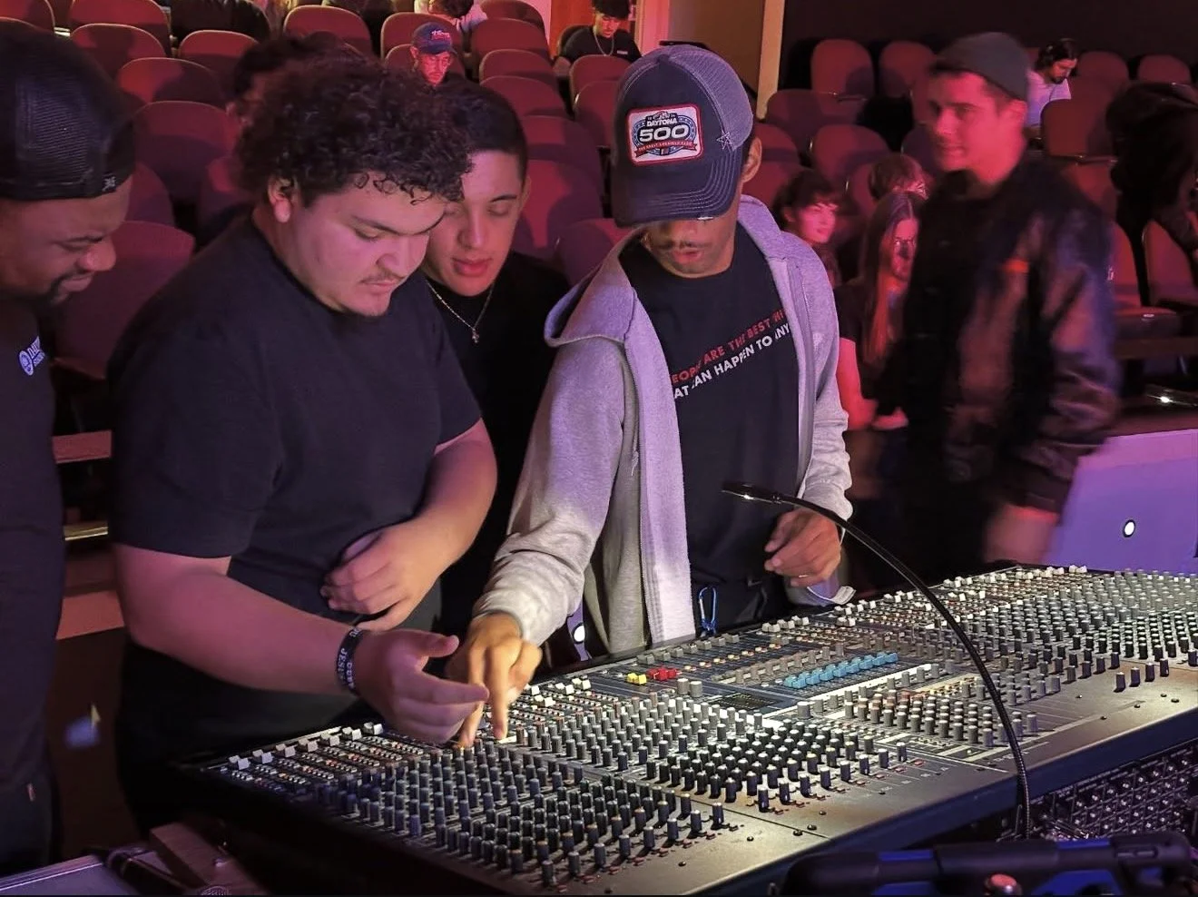 Several young people gather around a large audio mixing console with numerous knobs and sliders, some are adjusting controls while others observe attentively in a dimly lit room with purple and red lighting.