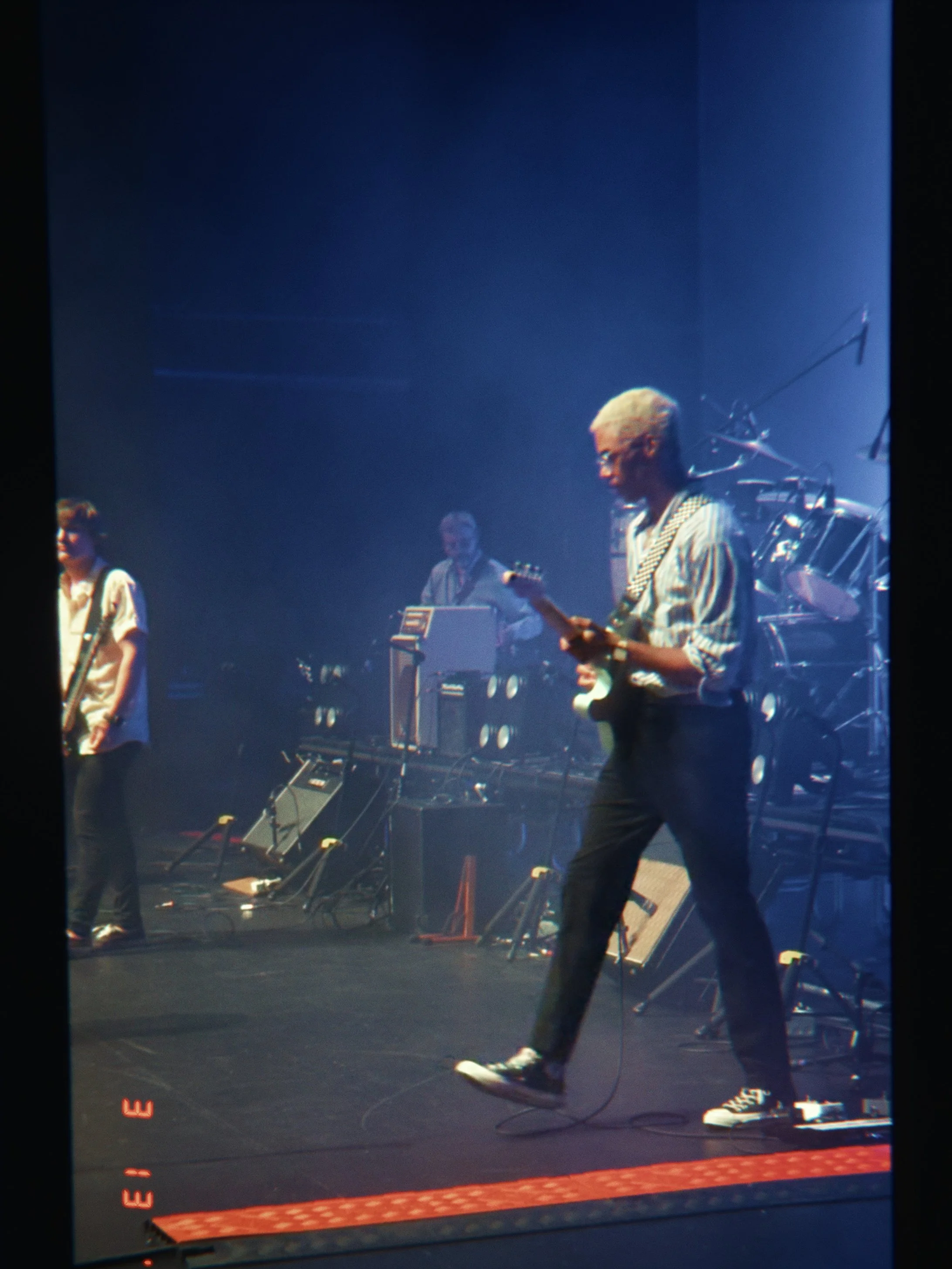 A musician with blonde hair wearing a striped shirt playing an electric guitar on stage, with band members and musical equipment in the background.