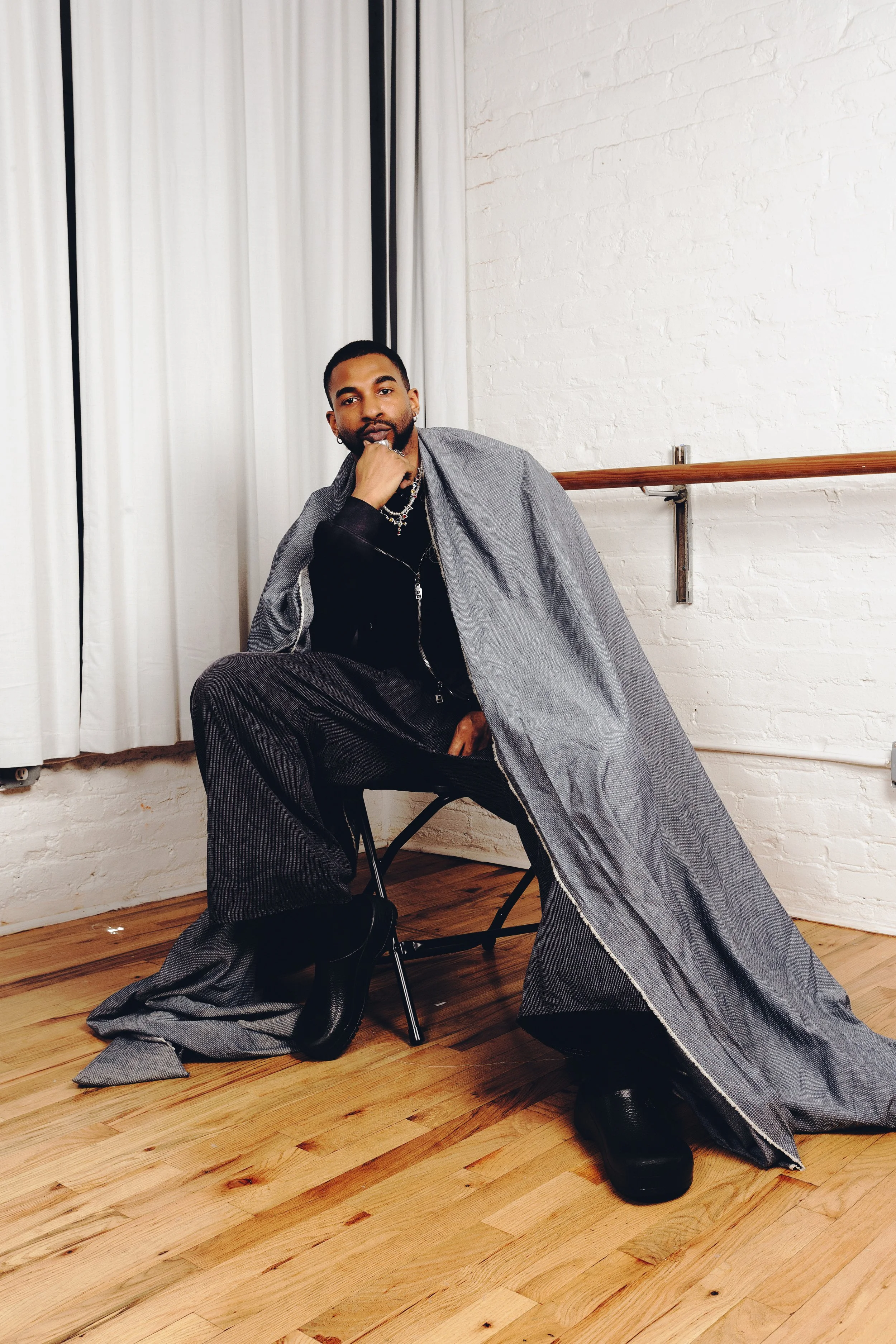 A man with dark hair and a beard, dressed in a black top and patterned pants, is seated on a chair in a room with white brick walls and wooden flooring. He has a gray coat draped over his shoulders and is wearing multiple necklaces. His pose is relaxed, with one hand resting near his chin and the other on his knee, looking directly at the camera.