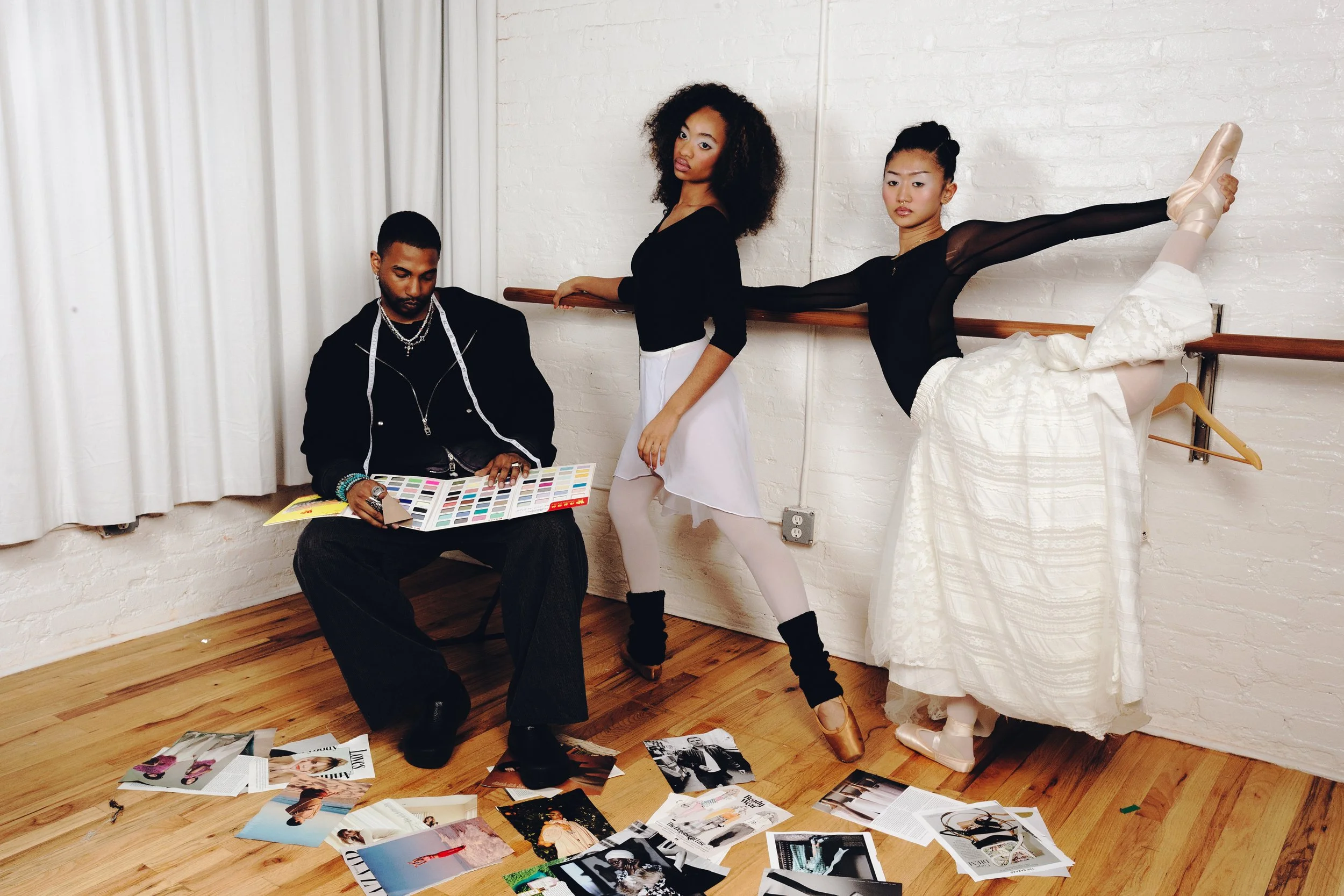 Three models in a studio setting, one sitting on a chair looking at magazine pages, one standing holding a ballet barre, and the third performing a ballet pose with one leg extended on the barre, surrounded by scattered magazines and photo spreads on the floor.