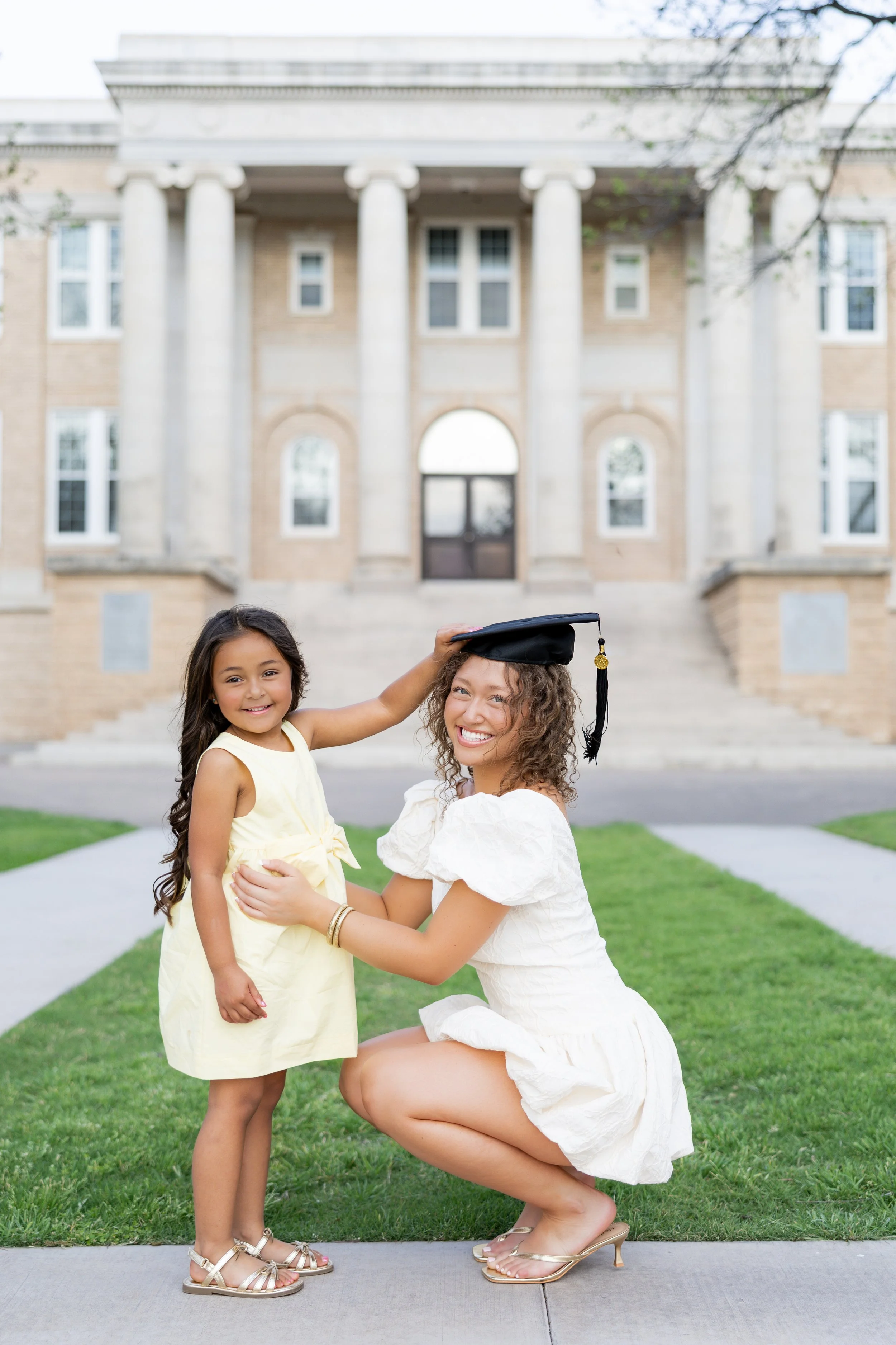 College graduate in a white dress posing with a young girl during an outdoor graduation session.