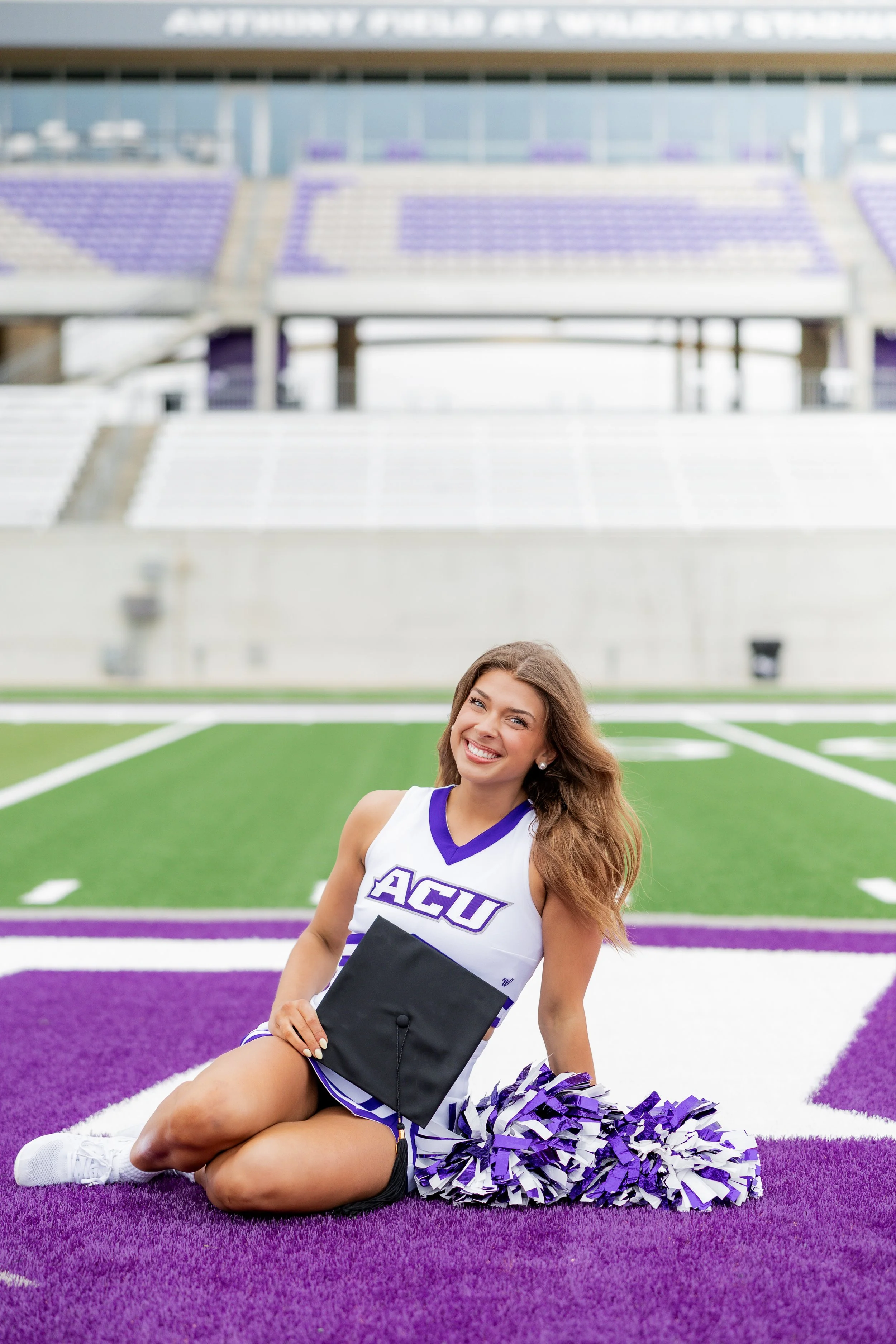 College cheerleader posing with her pom-poms on the football field during a graduation portrait session.