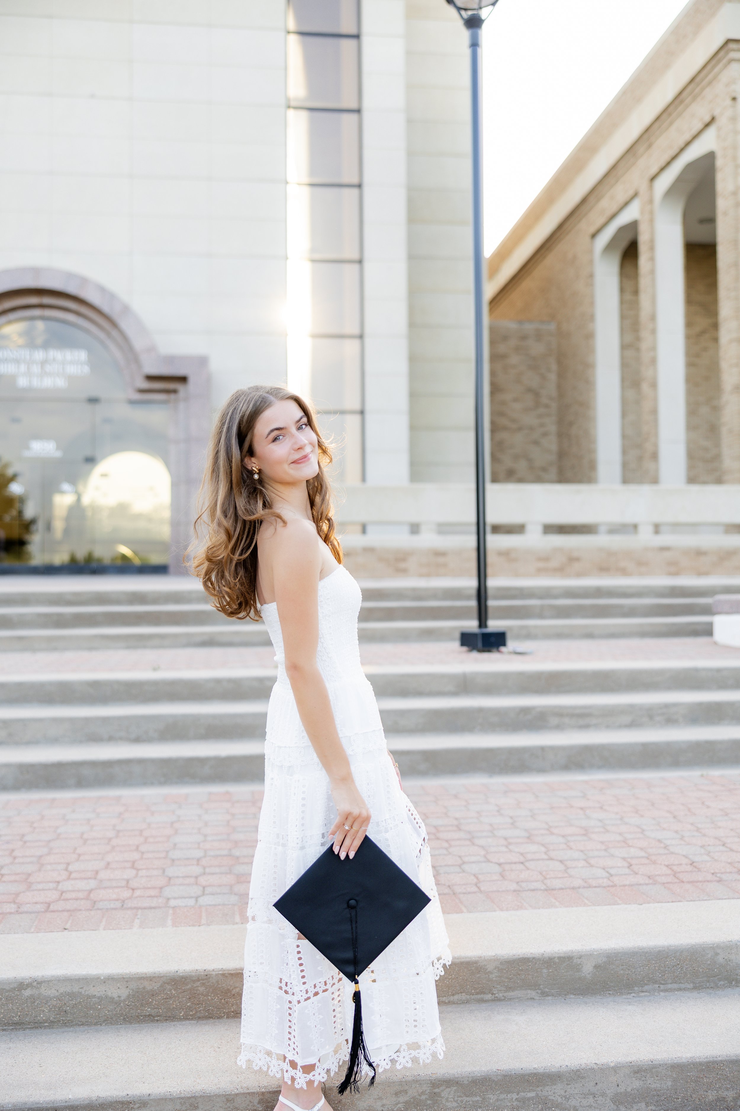 Graduate in a white dress holding her graduation cap while posing on outdoor steps.