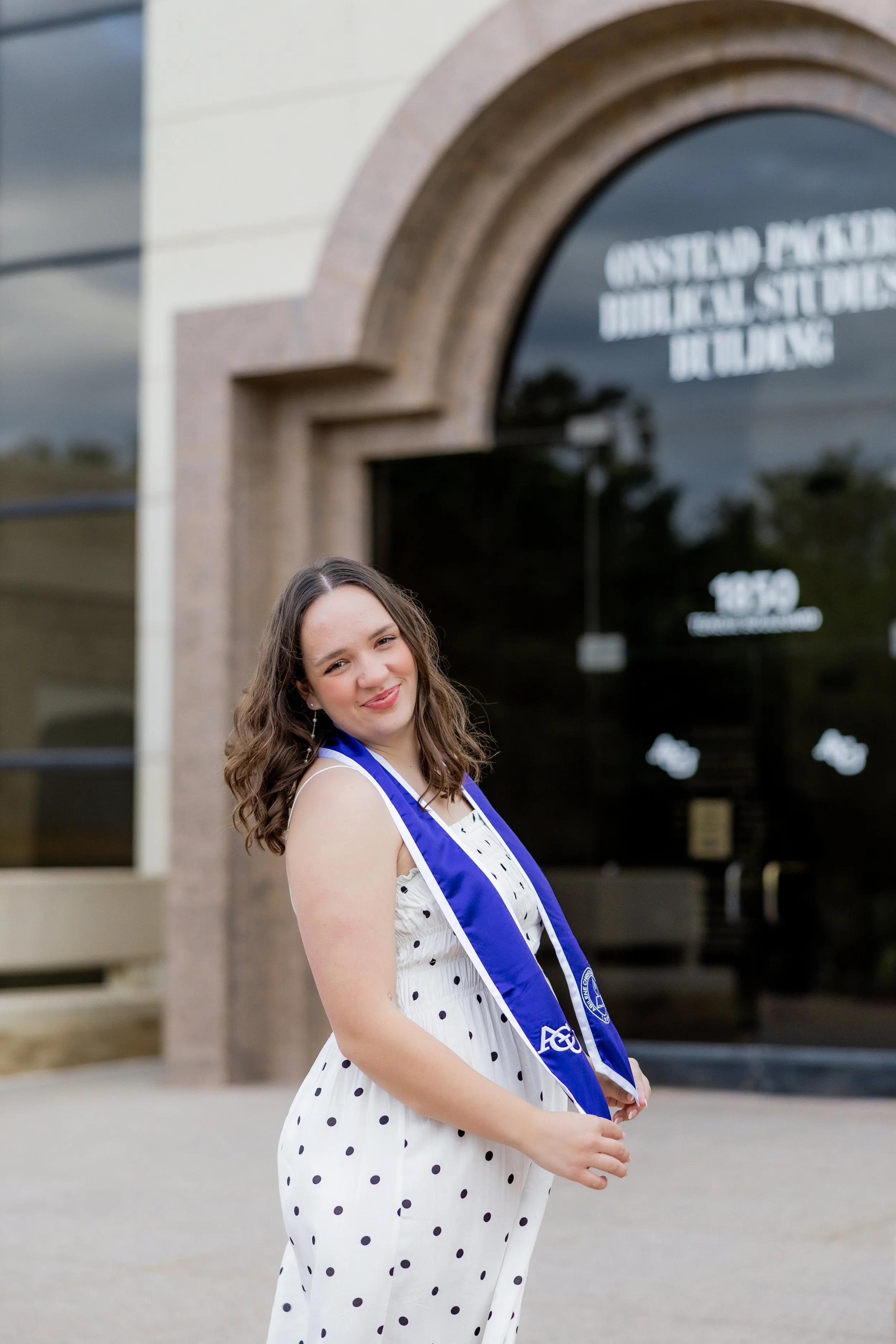 College graduate in a polka dot dress and graduation stole posing outside her university building.