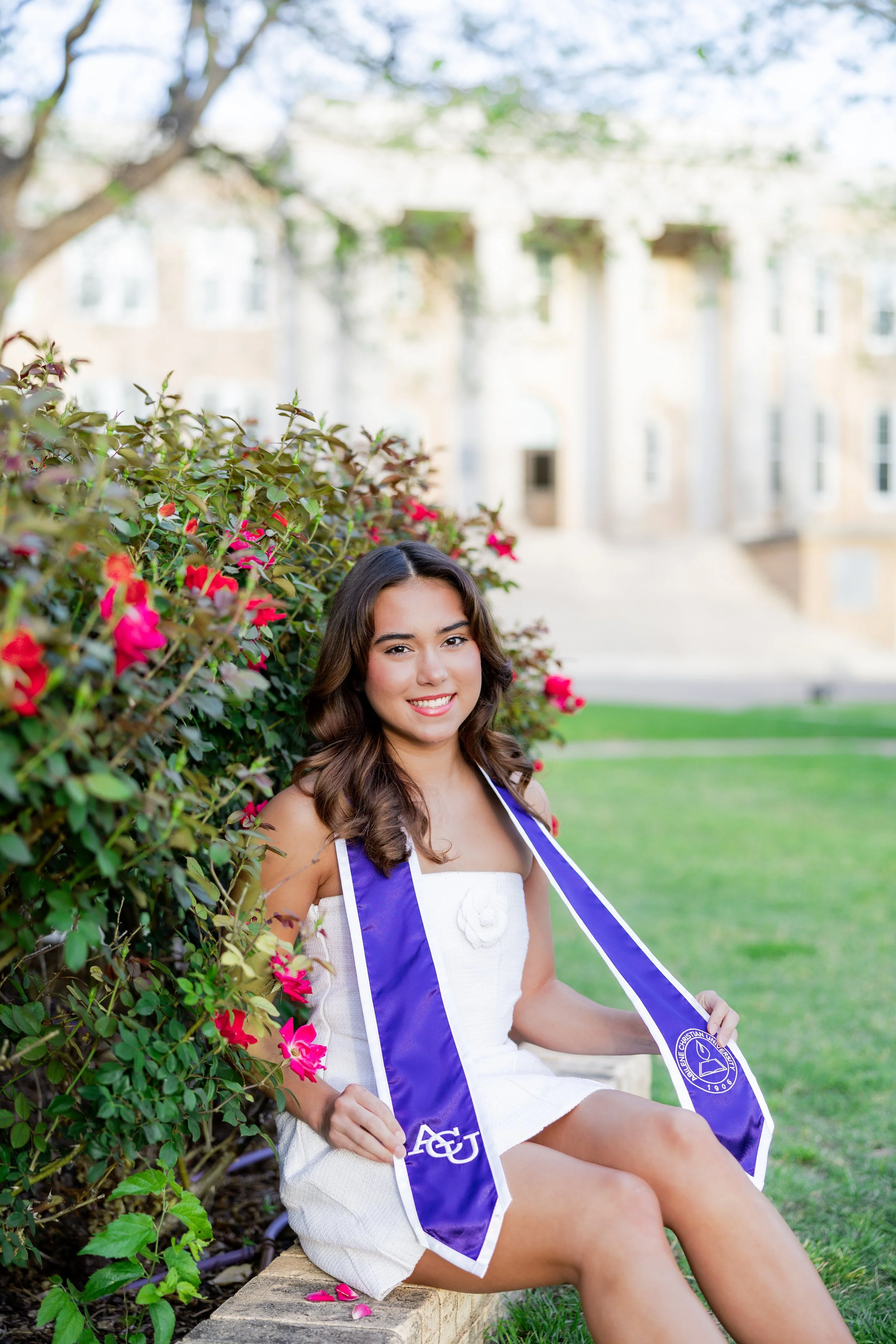 College graduate in a white dress and graduation stole posing outdoors surrounded by flowers during a graduation portrait session.