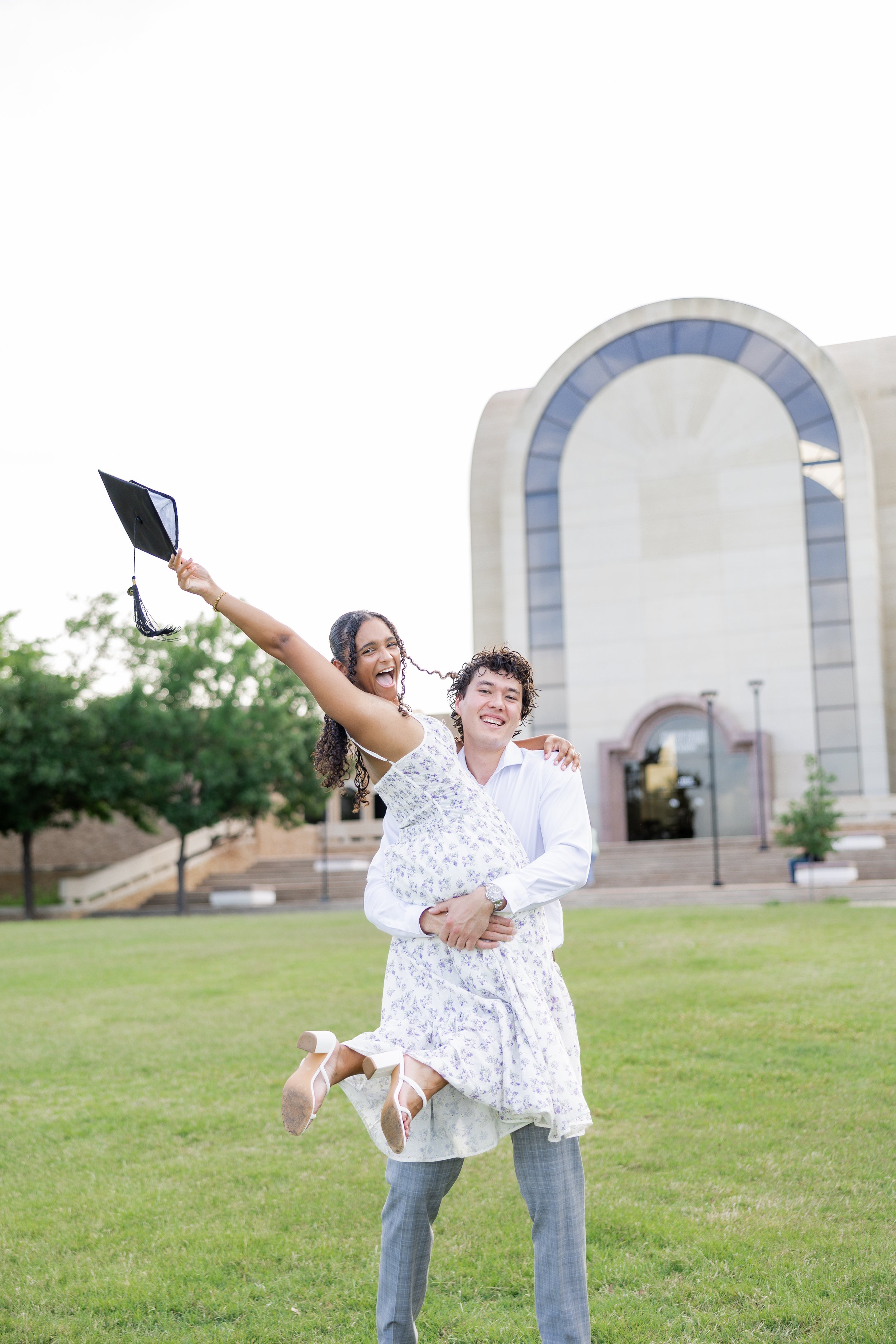 College graduate celebrating with her fiance while holding her graduation cap in the air.