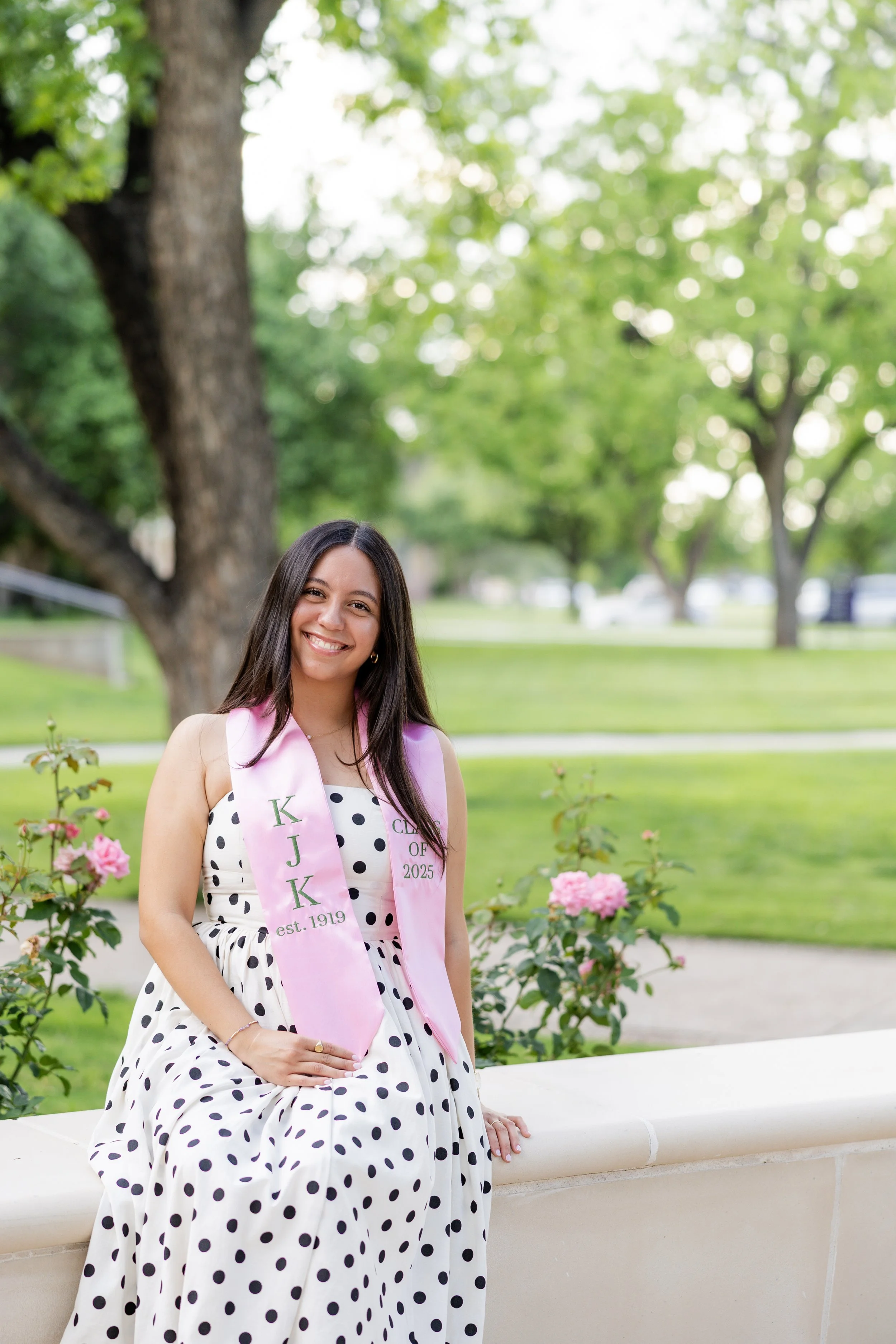 College graduate in a polka dot dress and graduation stole posing outdoors during a graduation photography session.