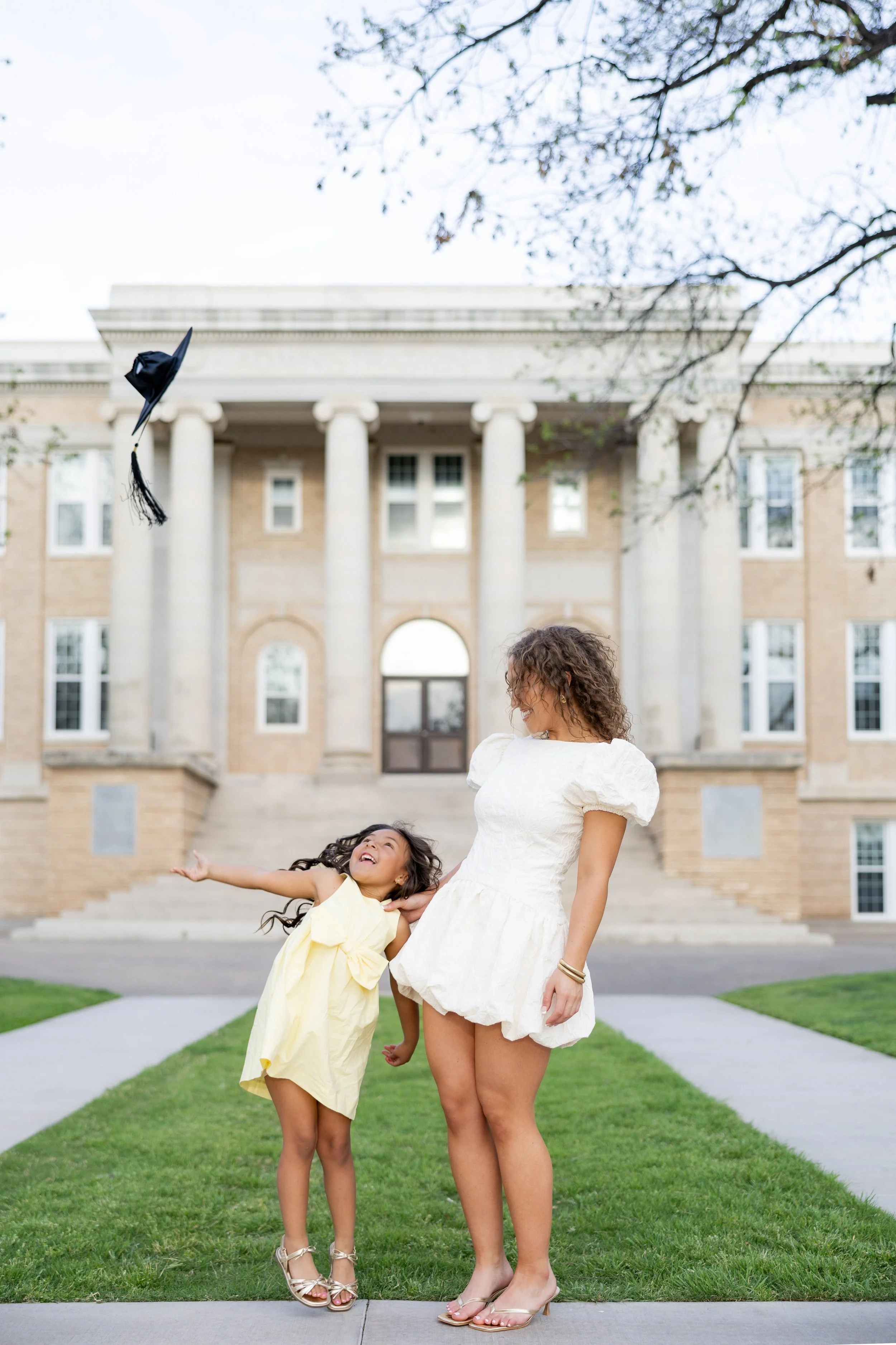 College graduate celebrating with her young daughter and tossing her graduation cap during an outdoor graduation session.