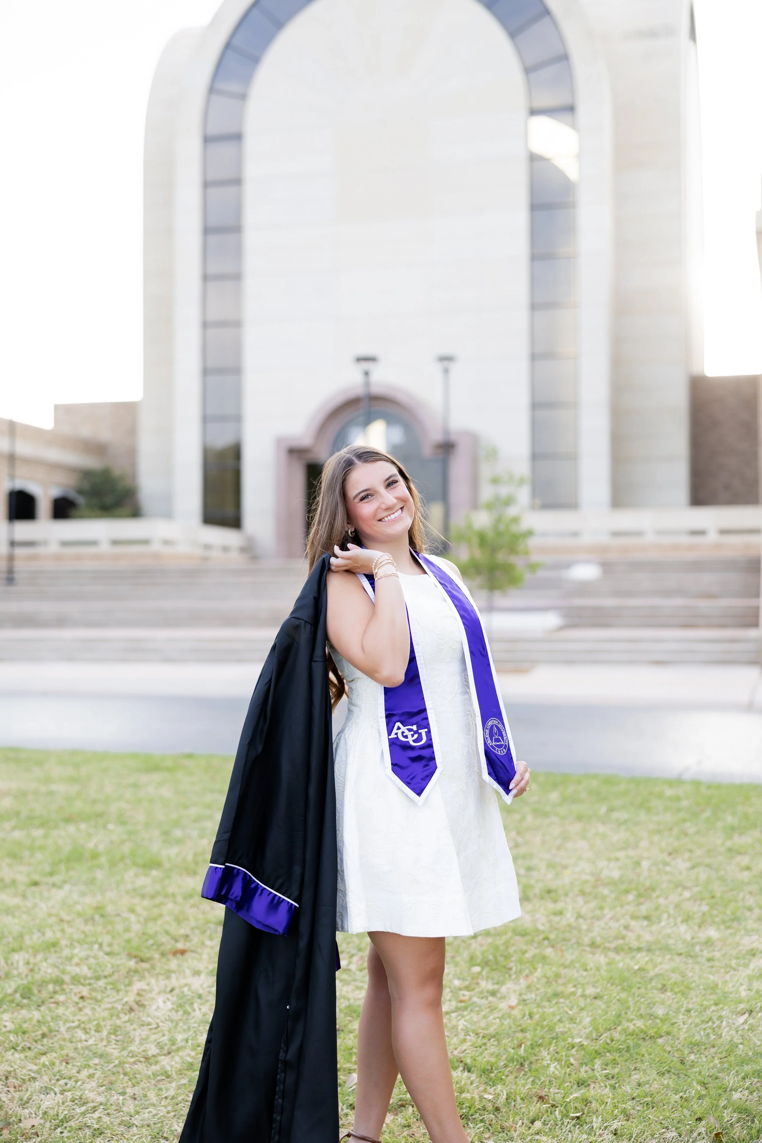 College graduate in her graduation gown and purple stole posing outdoors on campus.