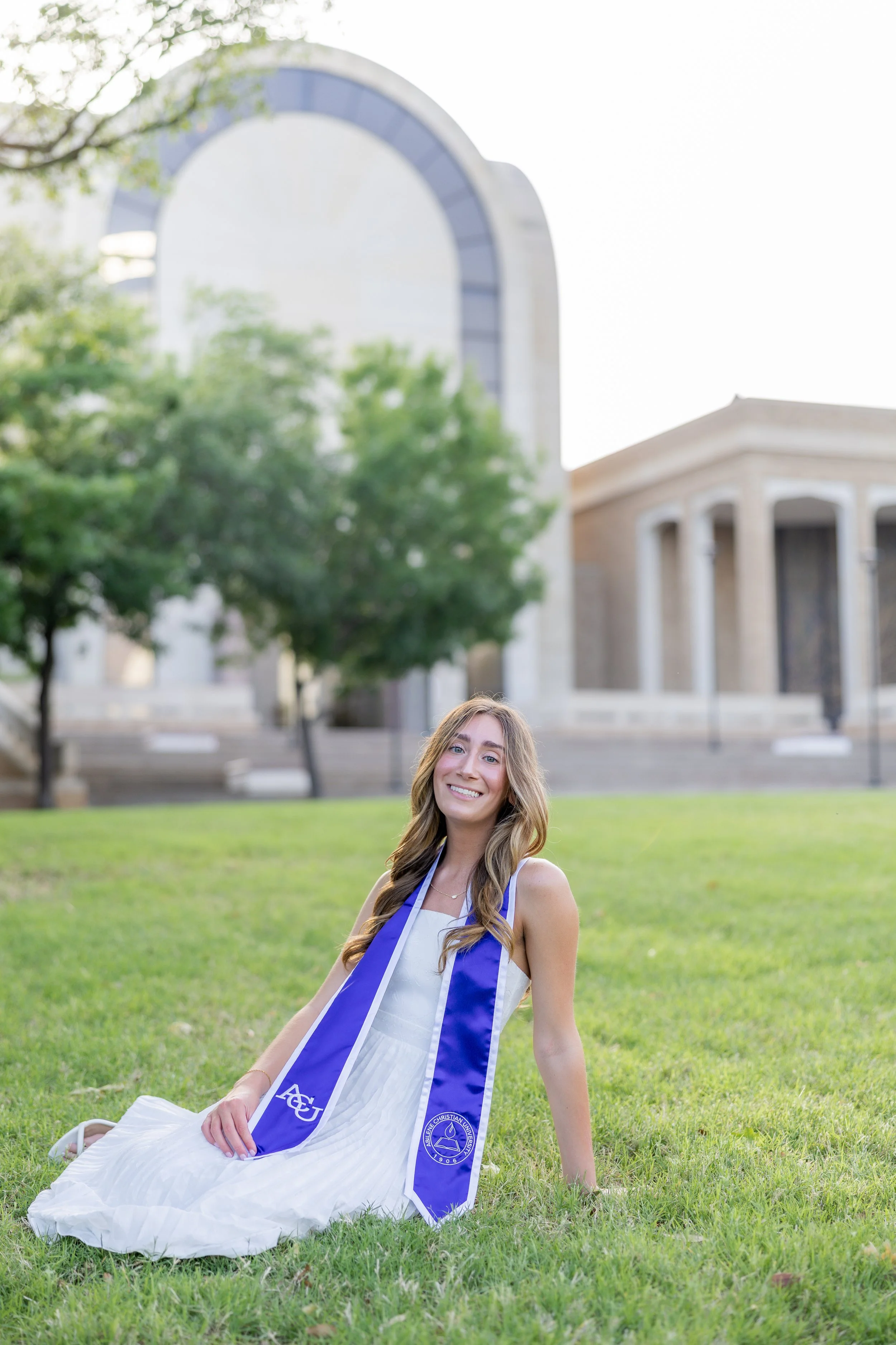 College graduate in a white dress and purple graduation stole posing on campus during a graduation session.
