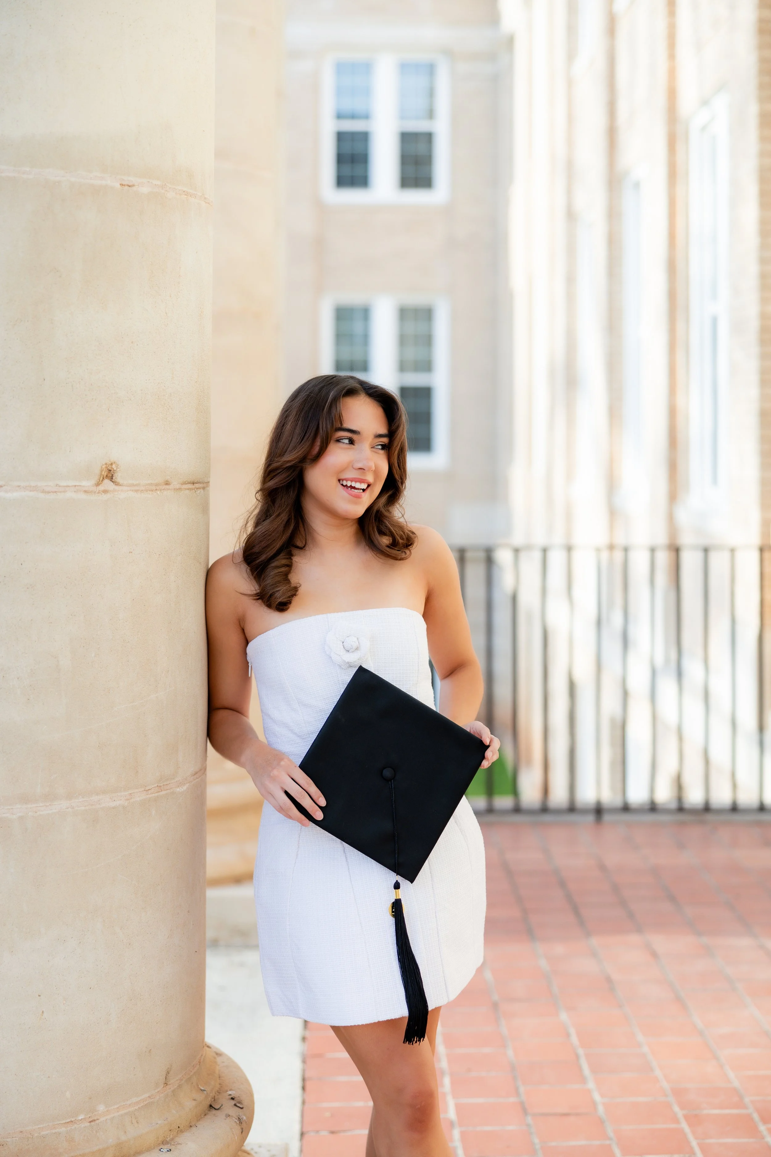 Graduate in a white dress holding her graduation cap during an outdoor graduation portrait session.