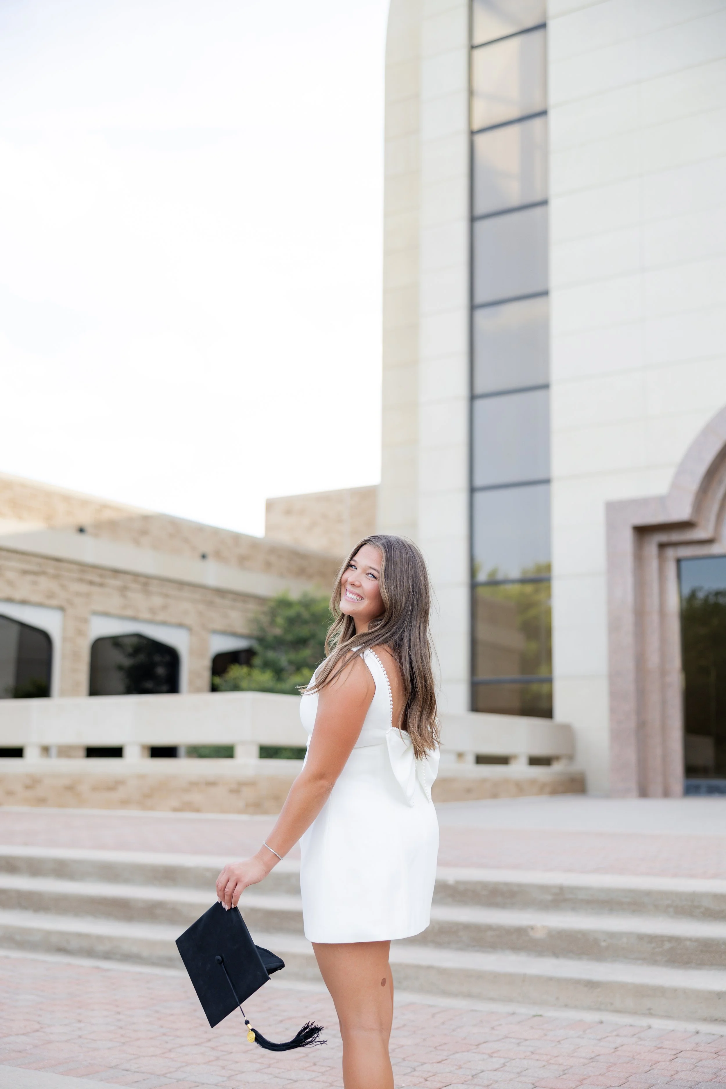 College graduate in a white dress holding her graduation cap during an outdoor graduation portrait session.