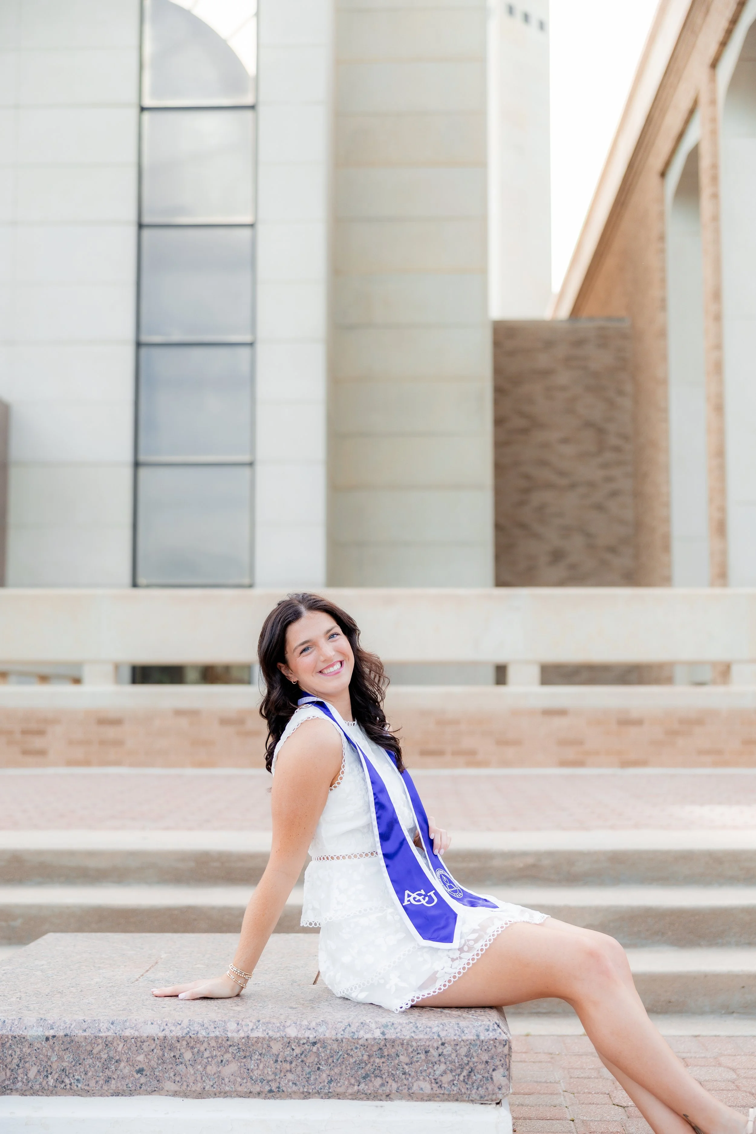 College graduate in a white dress and graduation stole posing on outdoor steps.