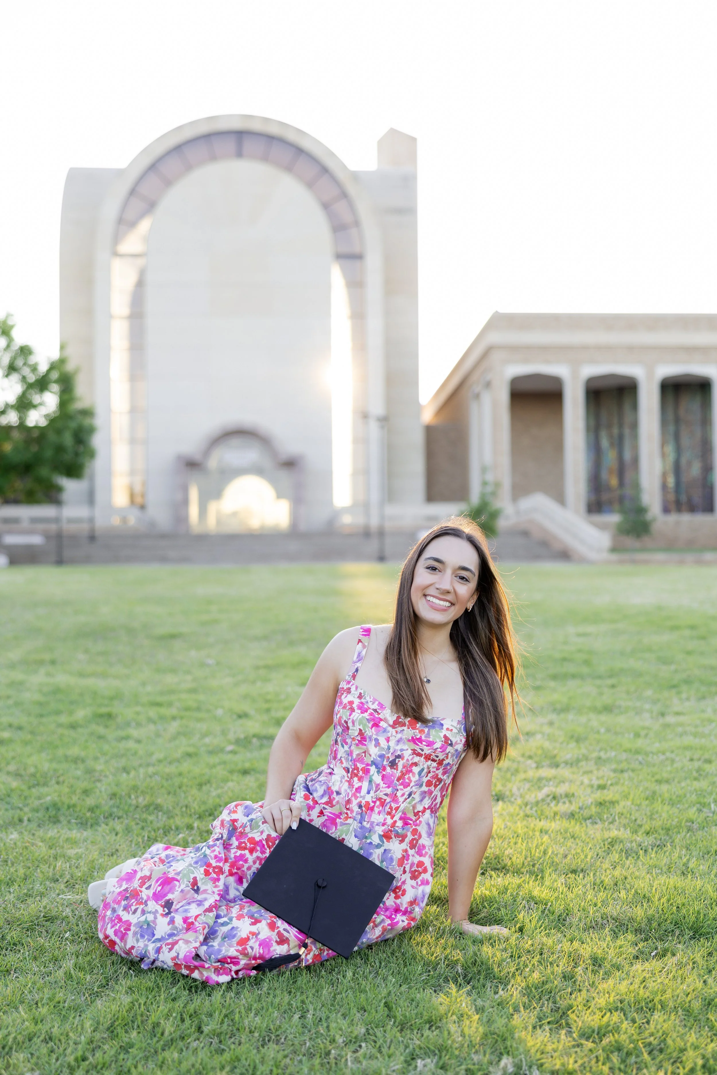 College graduate holding her graduation cap while sitting on campus at sunset.