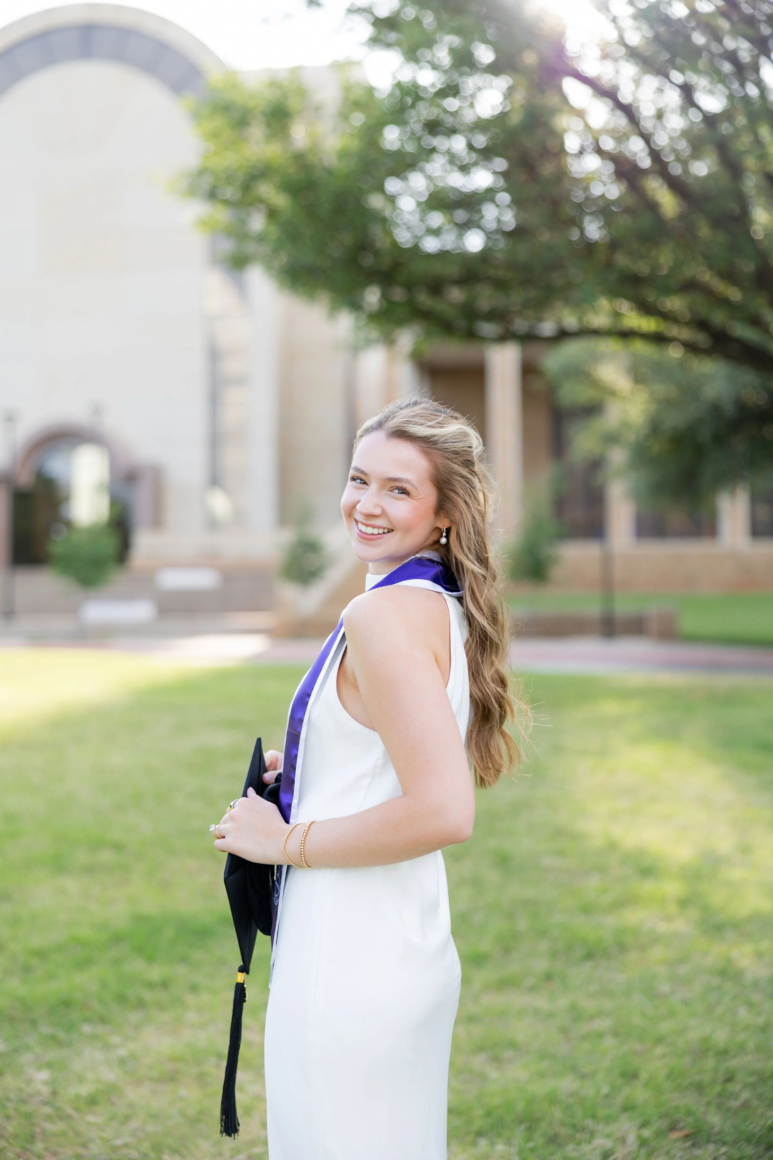 College graduate in a white dress holding her diploma during an outdoor graduation portrait session.